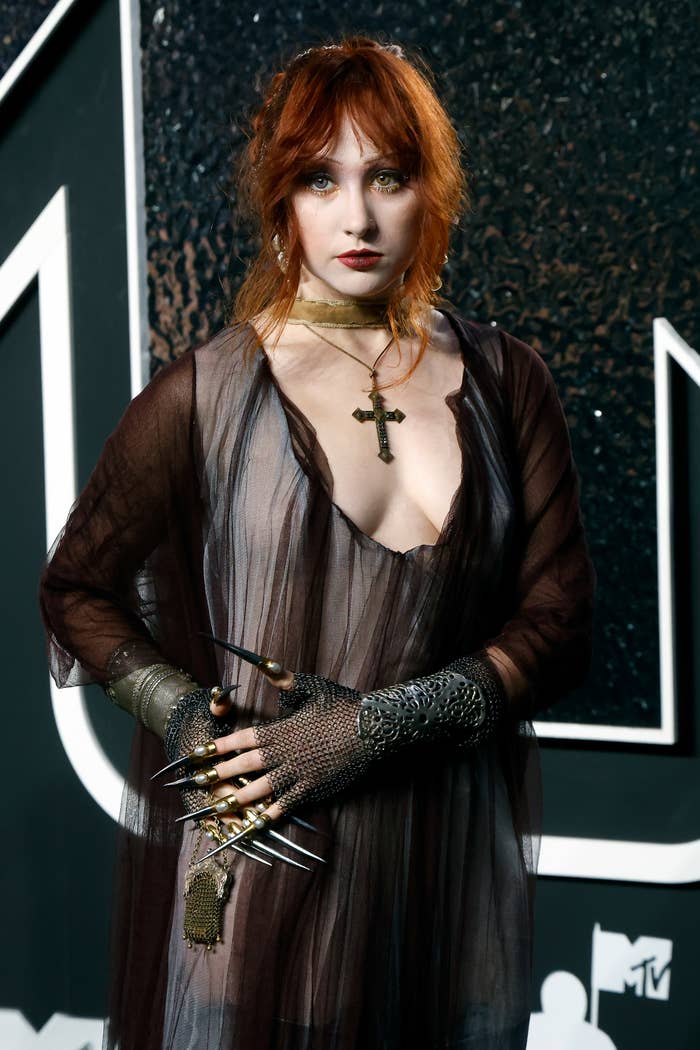 Chappell Roan at an event, wearing a sheer, flowing dress with a deep neckline, accessorized with a cross necklace and unique gloves with long metallic nails