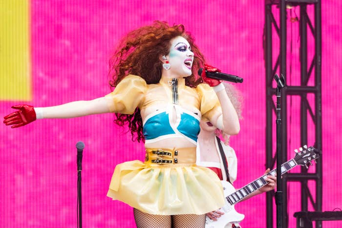 Drag performer in a futuristic costume sings and gestures on stage, with a band member playing guitar in the background
