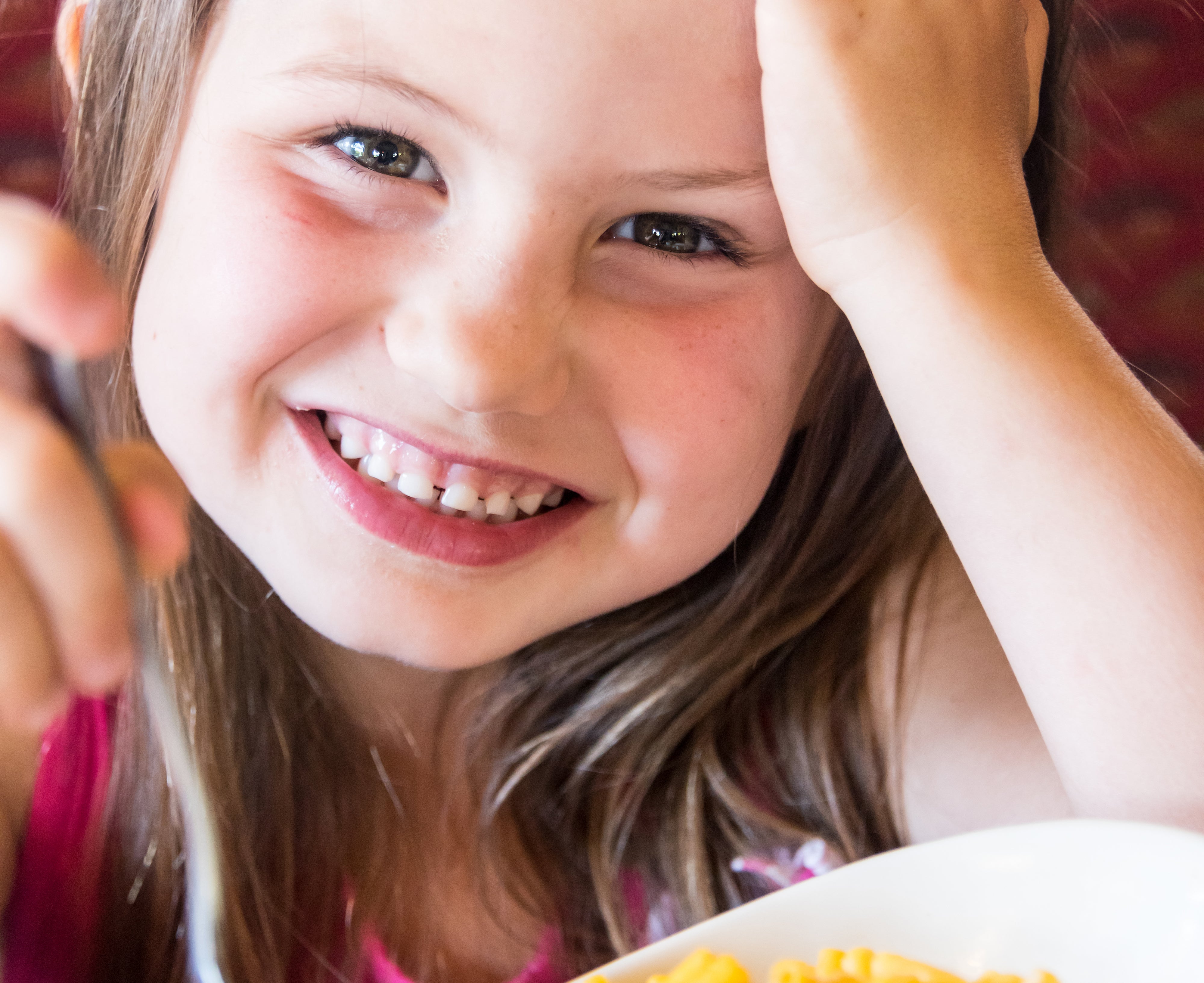 A smiling young girl is leaning her head on her hand while holding a spoon above a bowl of macaroni and cheese