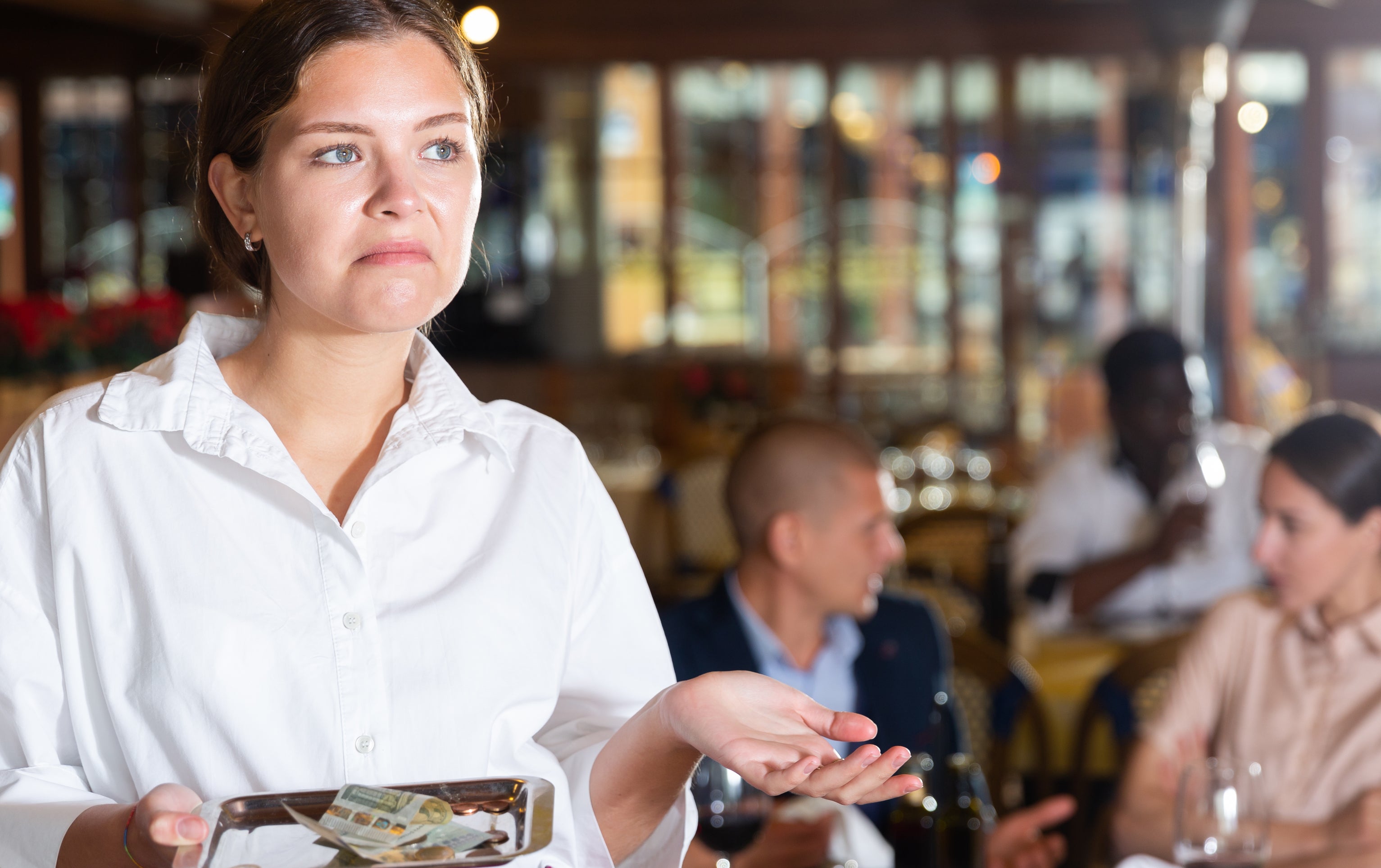 A restaurant server holds a plate with cash, appearing confused. In the background, diners are engaged in conversation