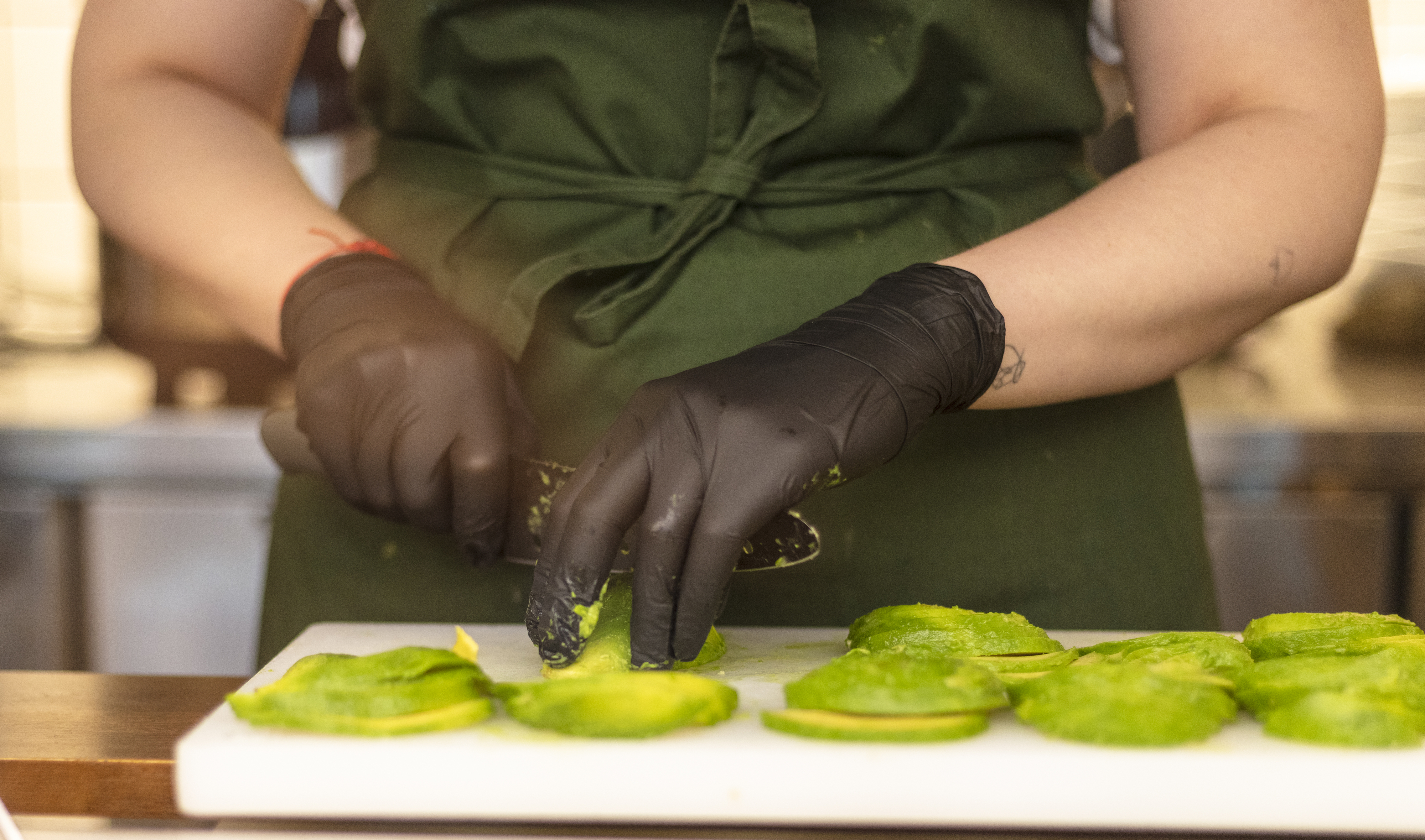 Person in a green apron and black gloves slices avocados on a white cutting board