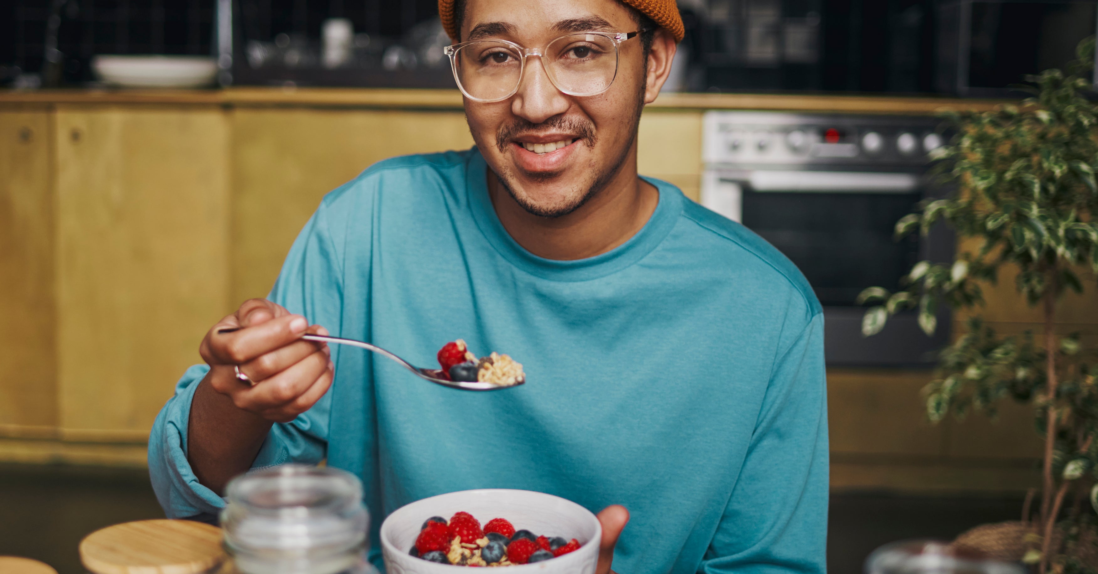 A person in glasses and a beanie smiles while holding a bowl of cereal with berries in a kitchen. Various jars and berries are on the table