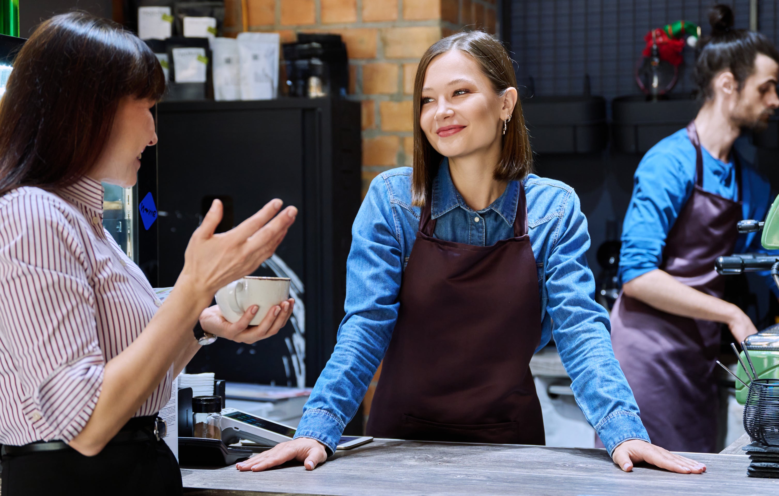 Two baristas, one man and one woman, behind a counter talking to a customer in a coffee shop. The man is making coffee, others are engaged in conversation