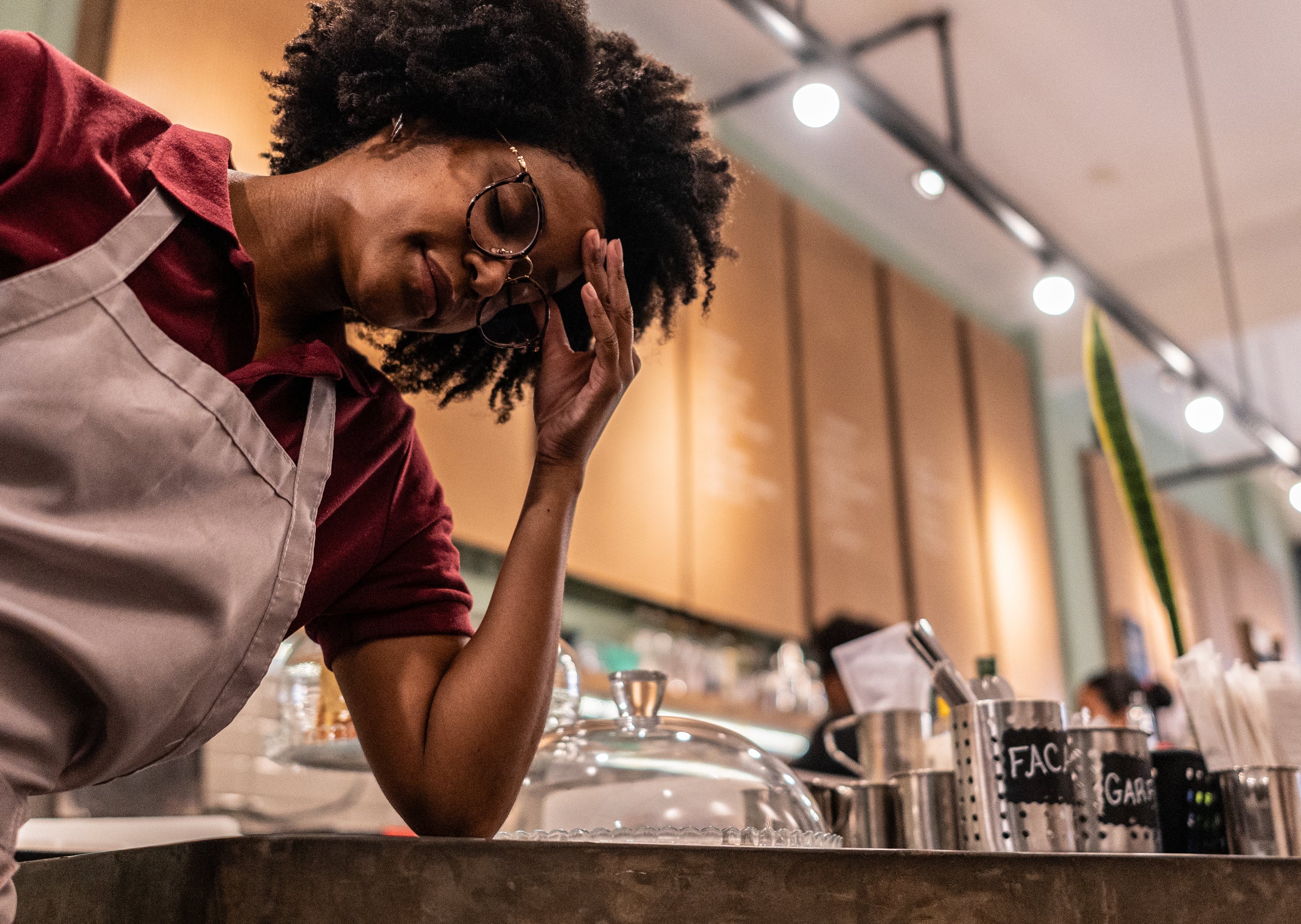 Person in an apron looks tired, leaning against a counter in a bustling café