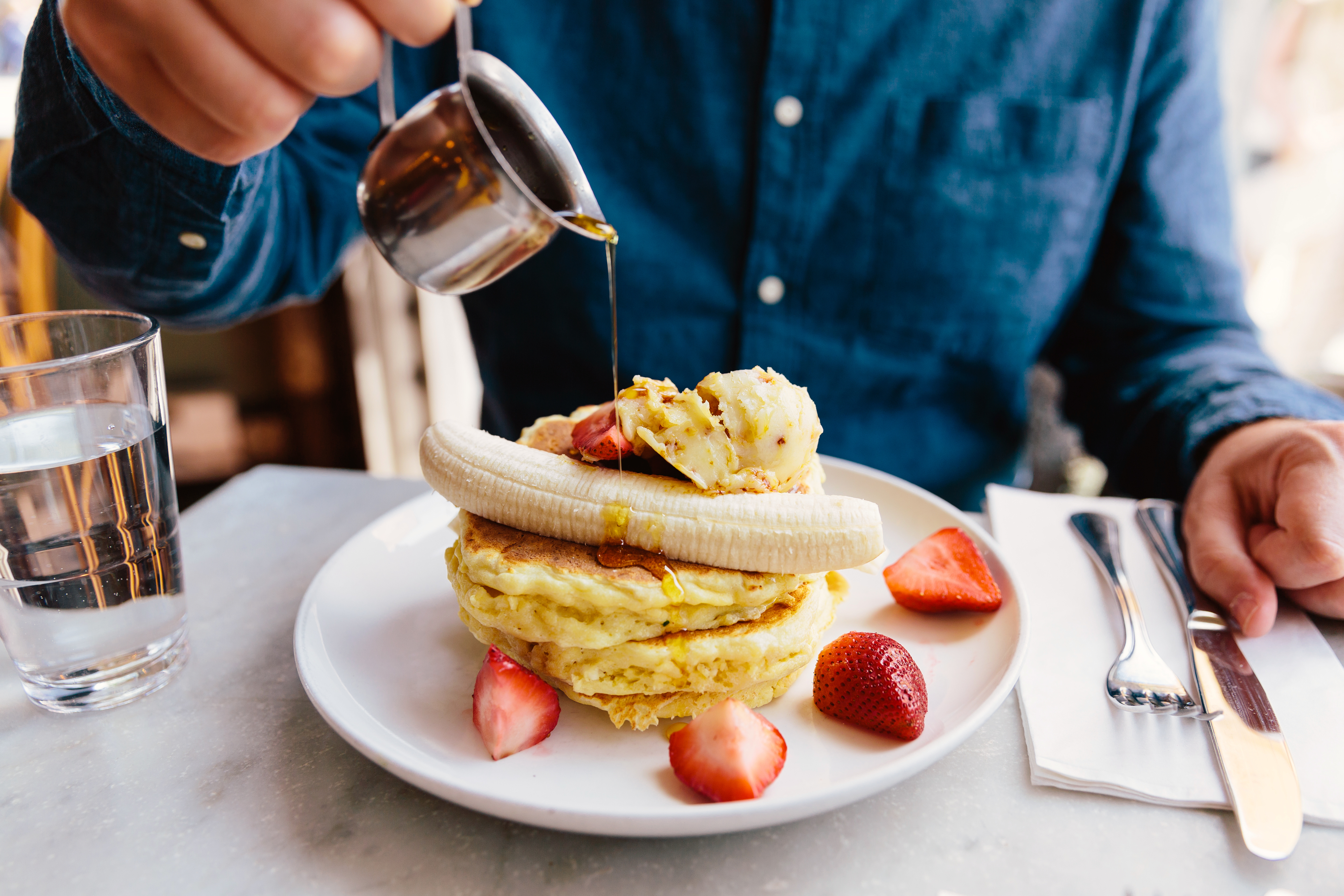 Close-up of a person's hand pouring syrup onto pancakes topped with banana slices and strawberries at a table with glass of water, fork and knife