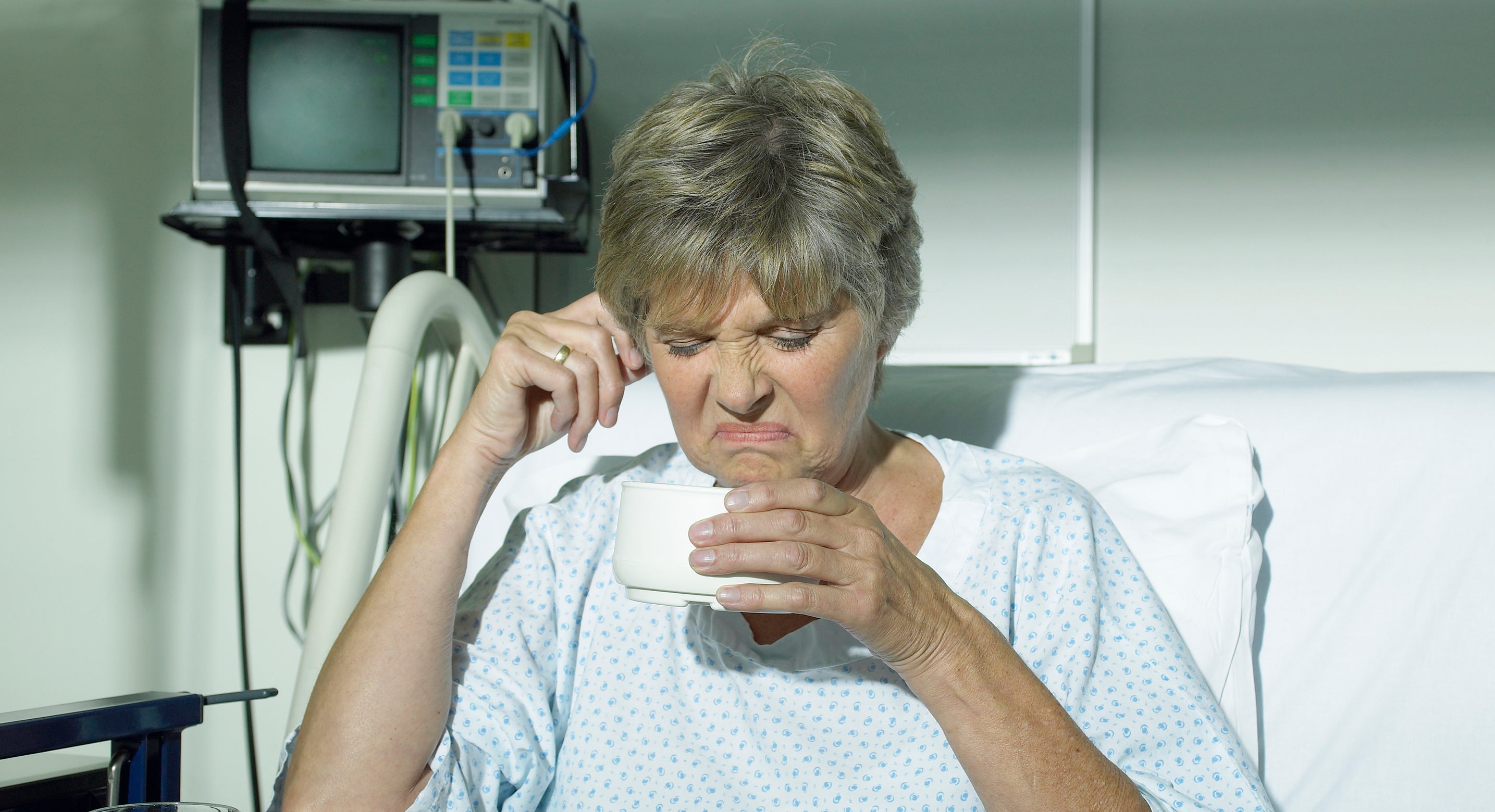 Elderly woman in a hospital bed, wearing a hospital gown, looking displeased while holding a bowl from her meal tray. Tray includes an apple, water, and mashed food