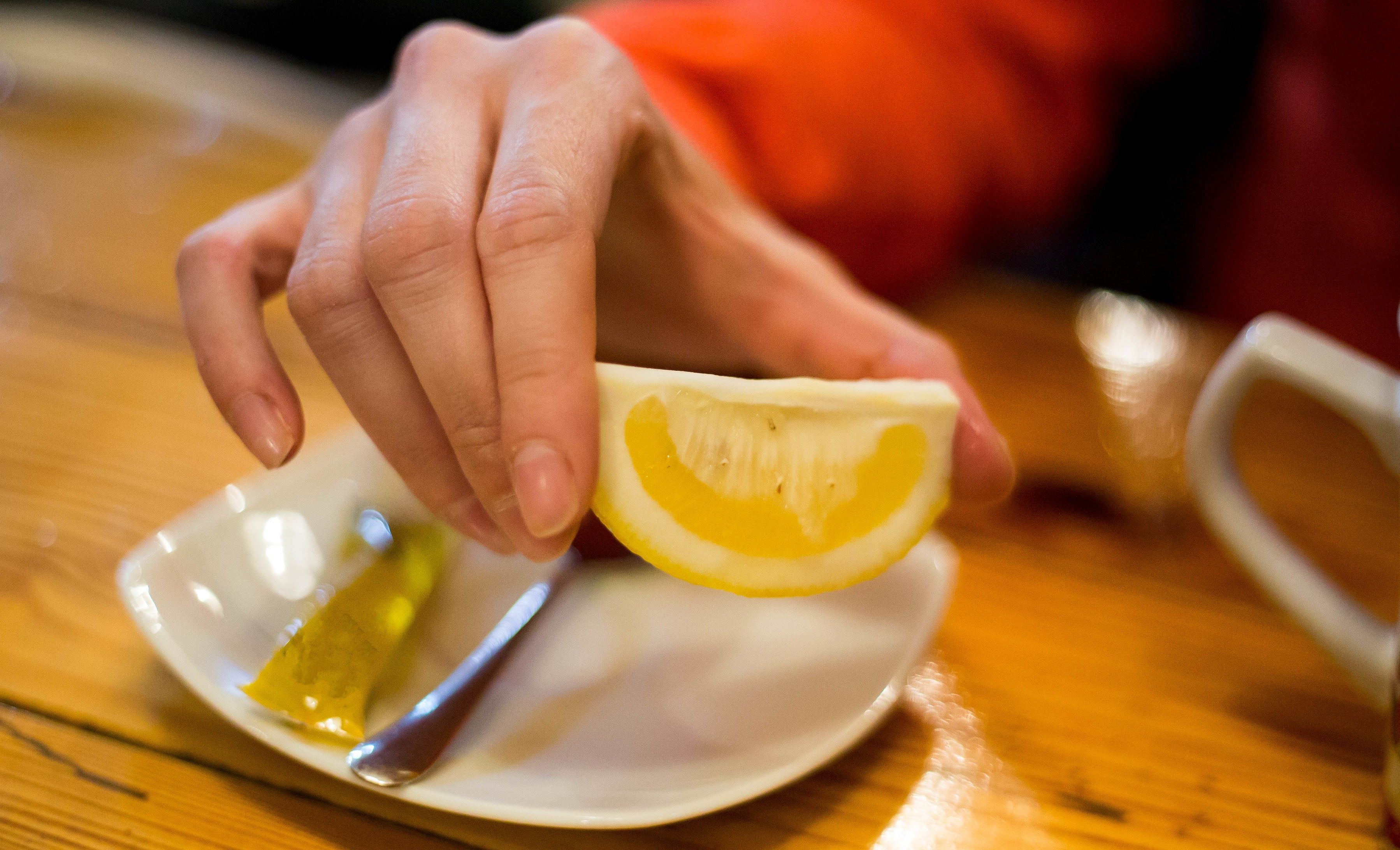 A hand holding a lemon wedge over a small plate with a pickle slice and a fork on a wooden table