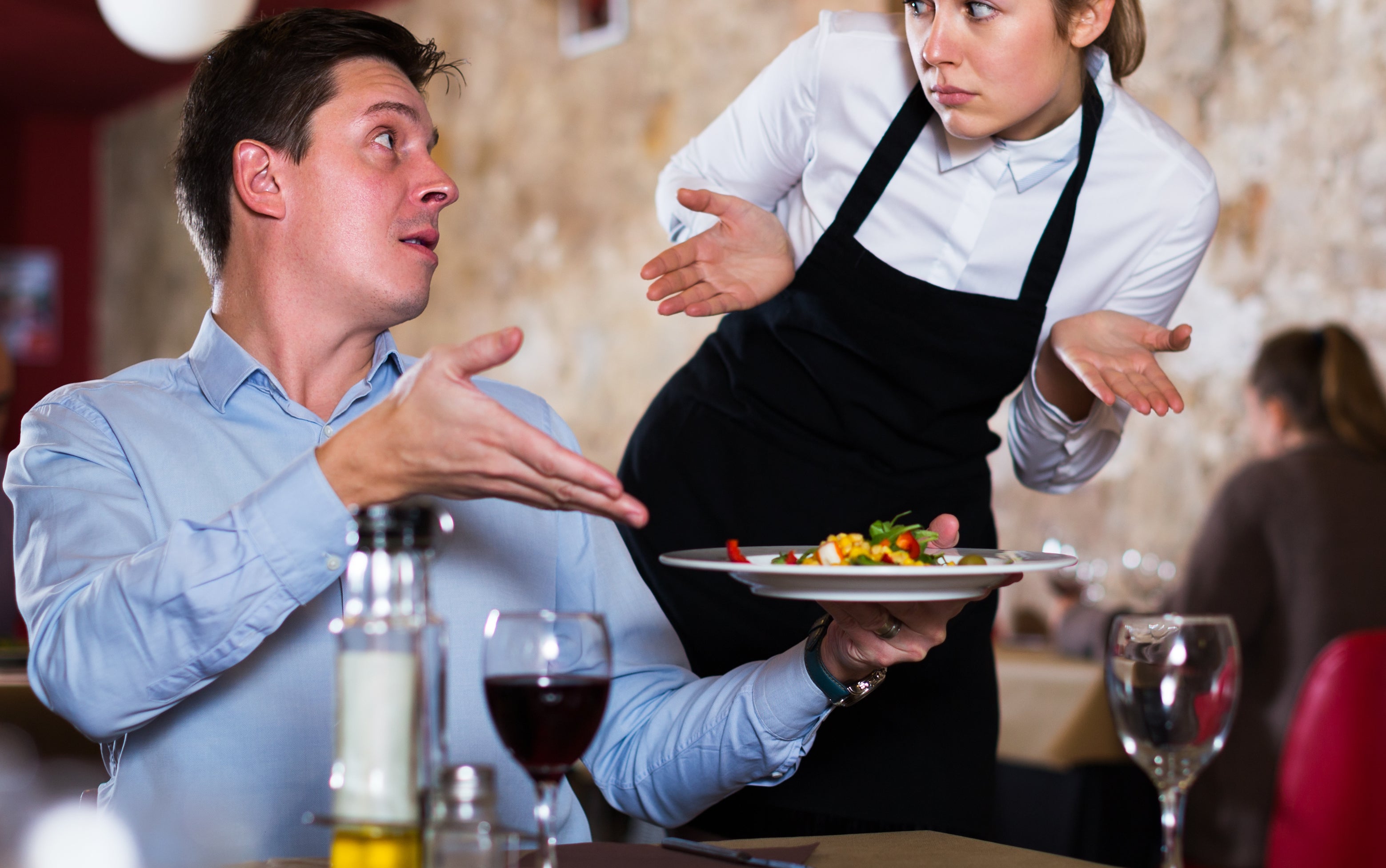 A server shrugs in confusion while a seated customer gestures with a plate of food. Others dine in the background