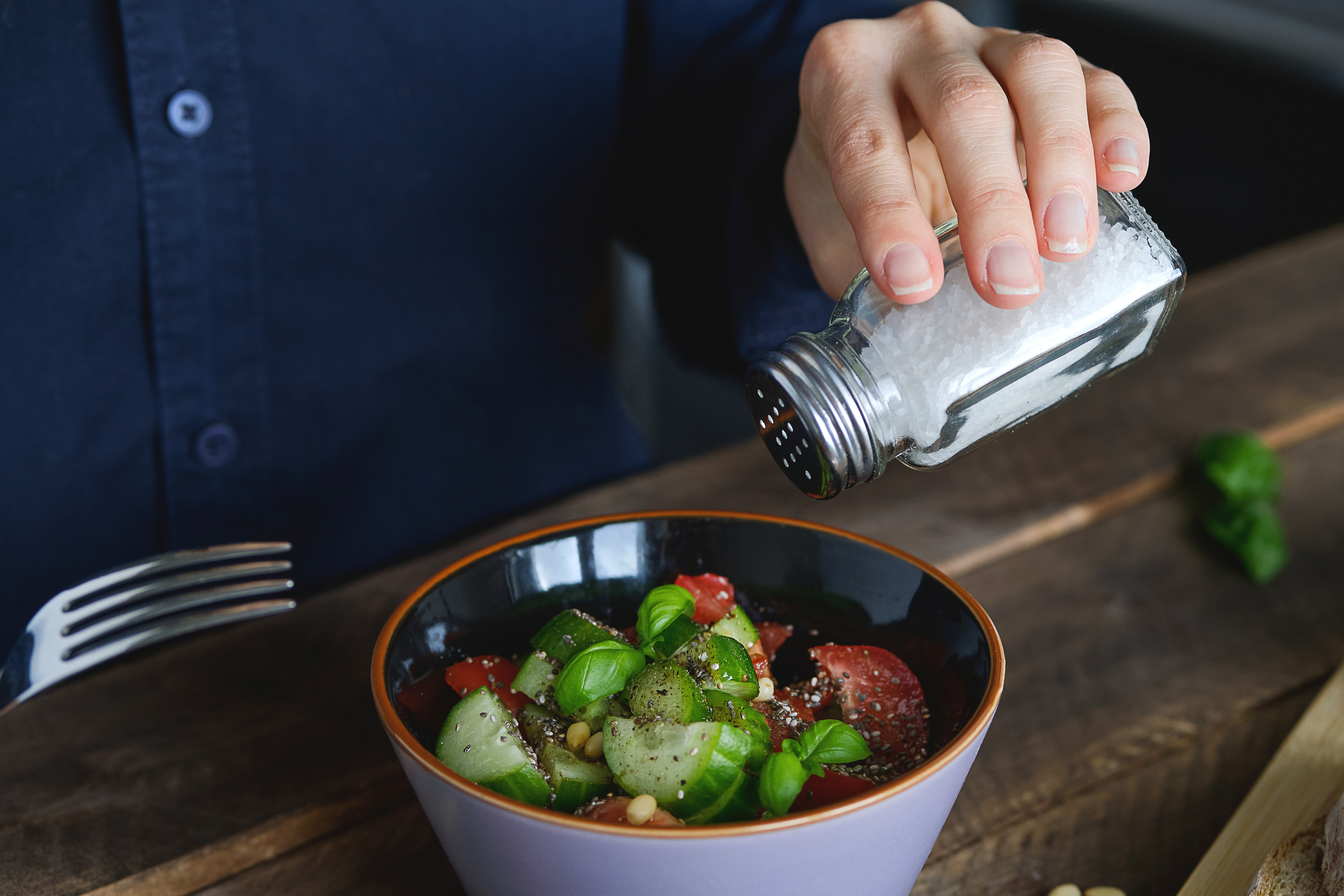 A person sprinkles salt over a bowl of fresh salad with cucumbers, tomatoes, and basil on a wooden table