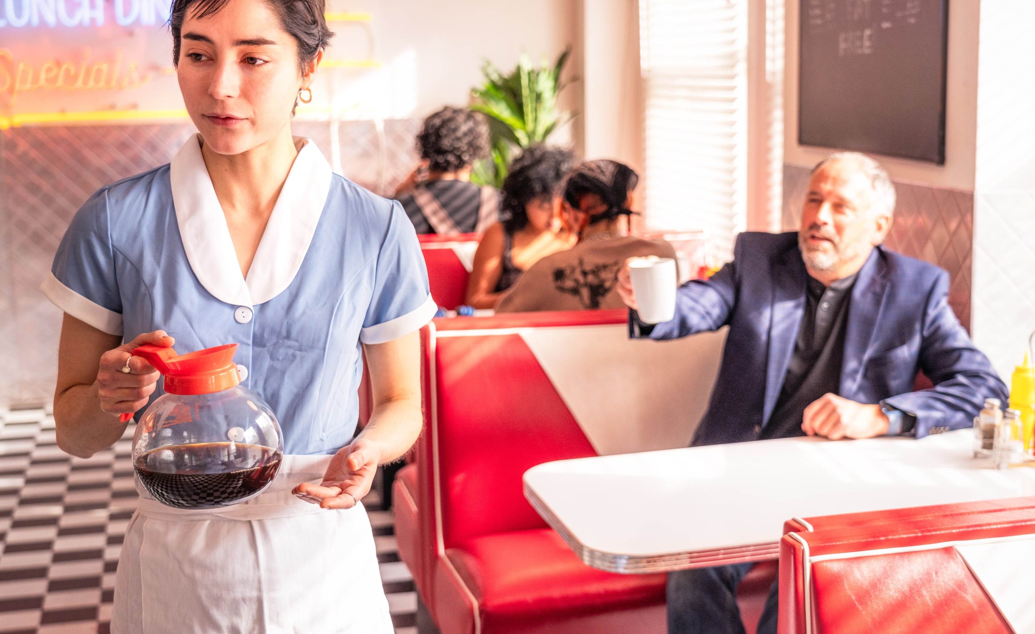 A waitress carries a pot of coffee in a diner, while a man in a suit sits in a booth. Two other people are seated in another booth in the background