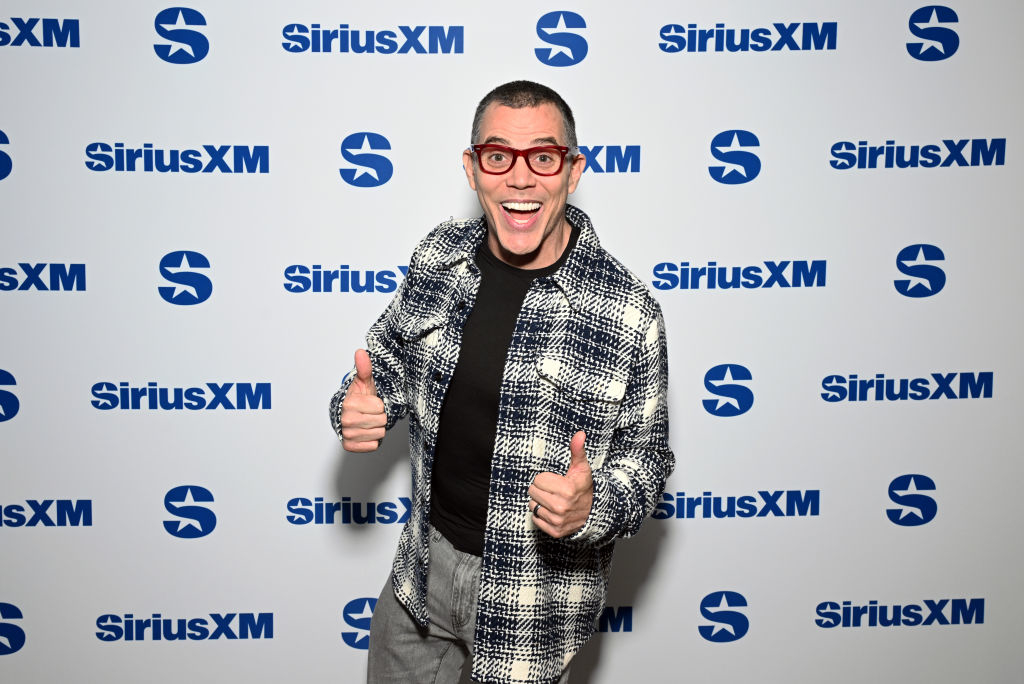 Steve-O smiling, wearing red glasses, a plaid jacket, and giving two thumbs up in front of a SiriusXM backdrop