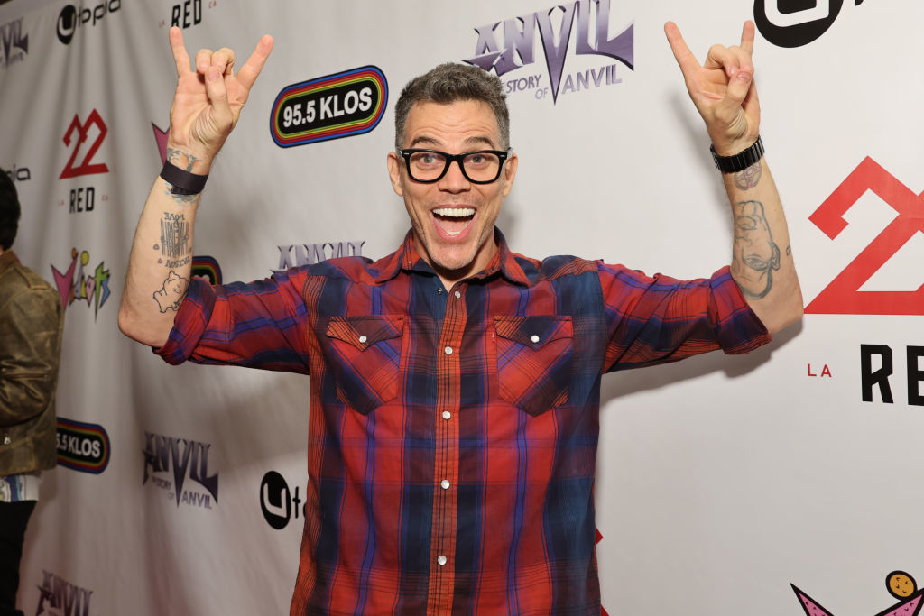 Steve-O poses on a red carpet event, wearing a plaid shirt, holding up rock-and-roll hand signs