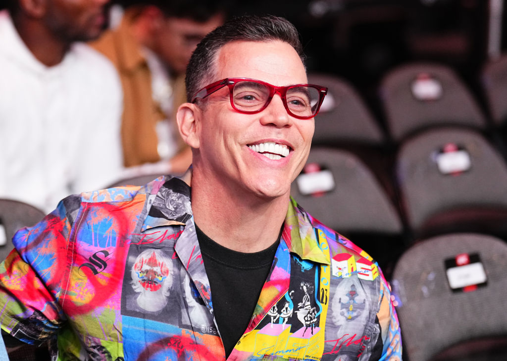 Steve-O smiles wearing a vibrant, patterned shirt and red glasses at an event