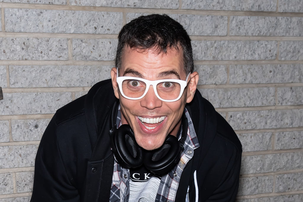 Steve-O smiles widely while wearing glasses, a checkered shirt, and a hoodie with headphones around his neck