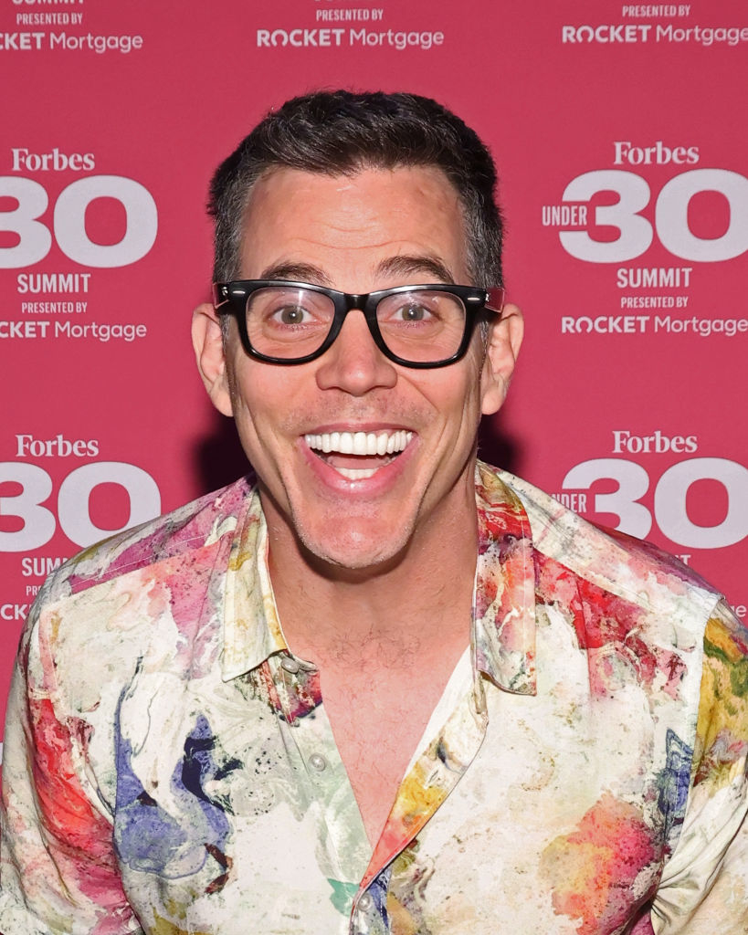 Steve-O at Forbes Under 30 Summit event, wearing glasses and a unique, colorful shirt with a paint-splatter design, smiling broadly