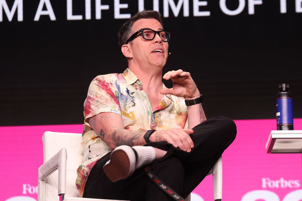 Steve-O speaks at a Forbes event, sitting on a panel chair, wearing a casual floral shirt and glasses. A portion of a text banner is visible behind him