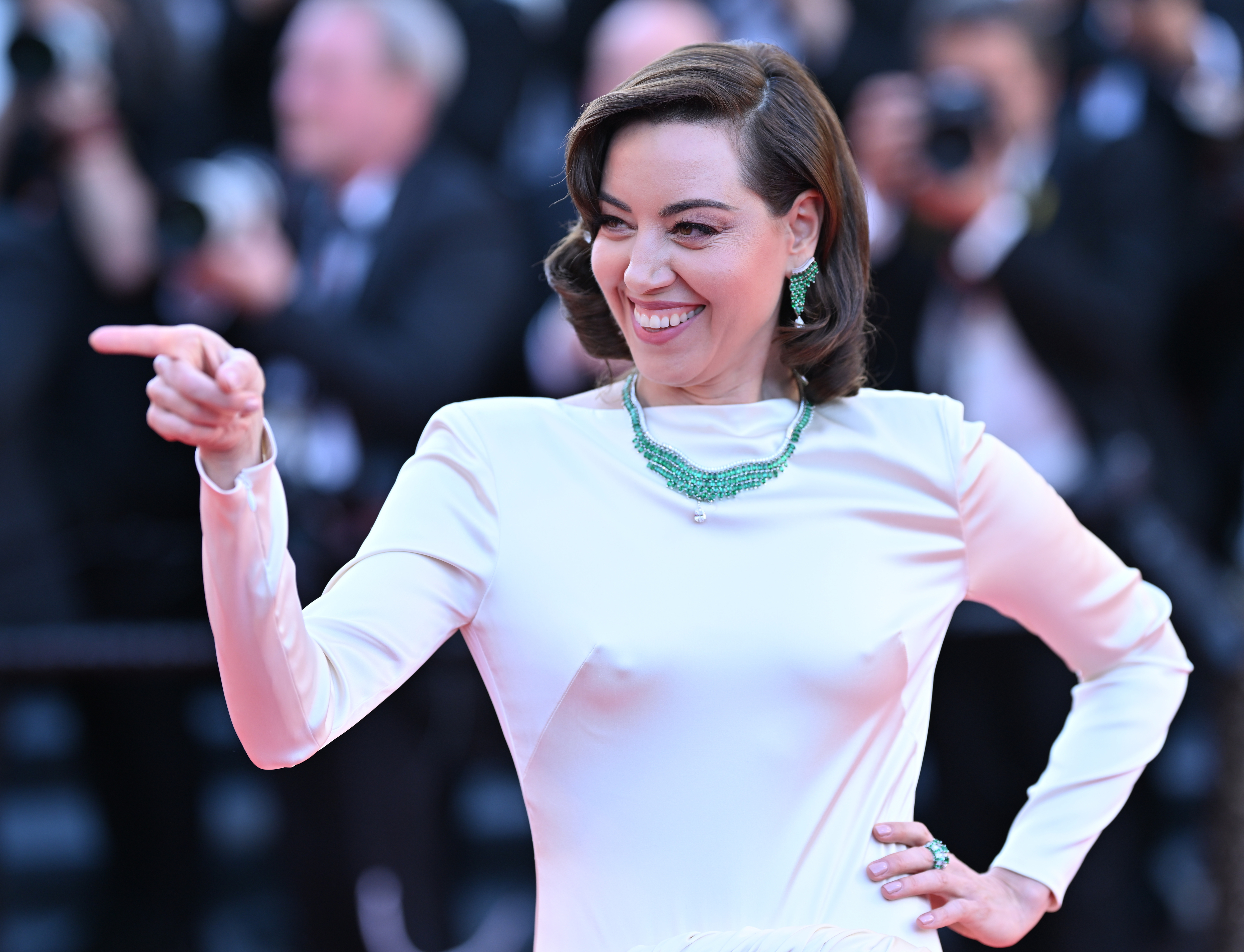 Aubrey Plaza on the red carpet wearing a sleek, long-sleeve gown with emerald green jewelry, smiling, and pointing to the side