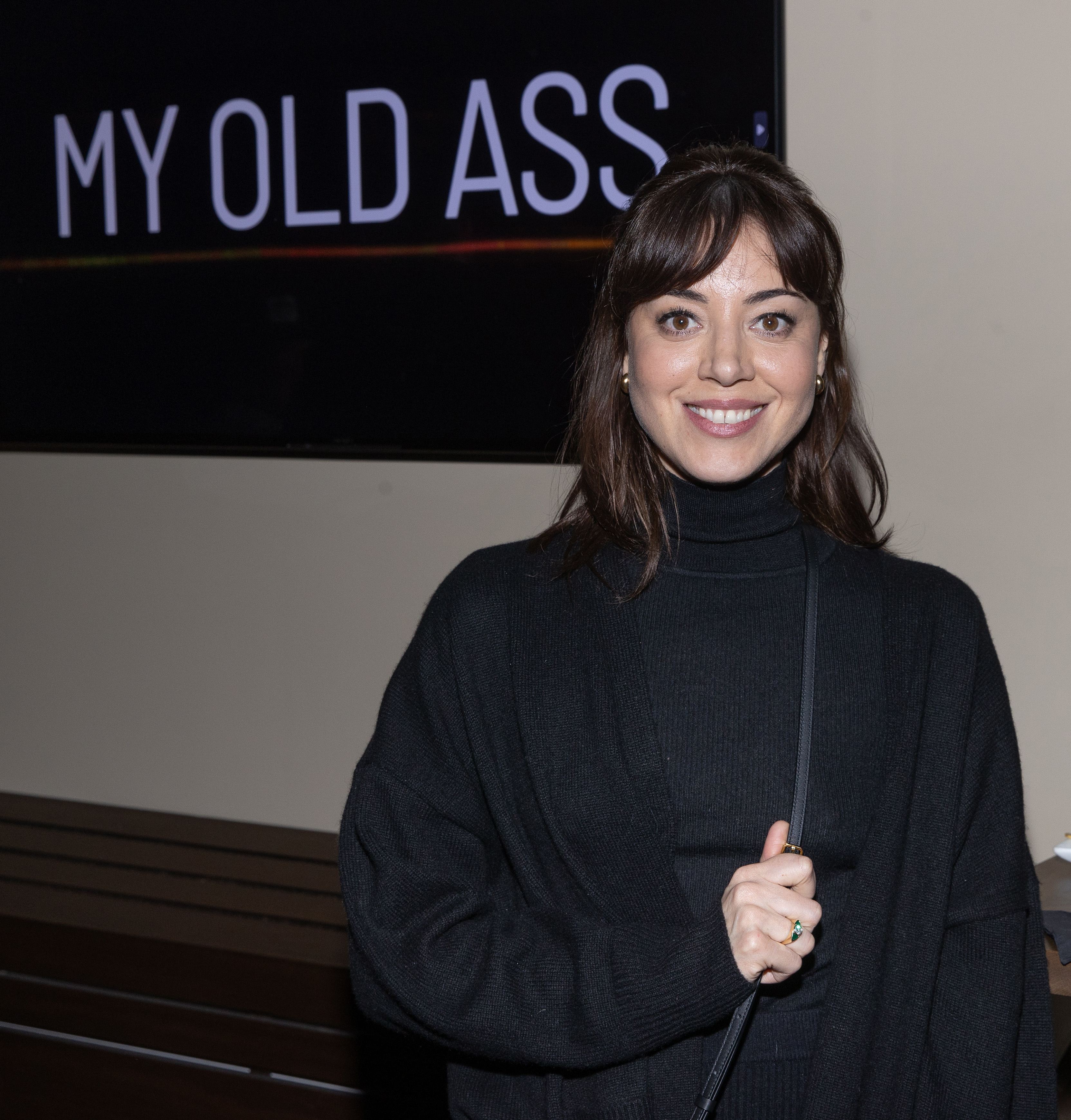 Aubrey Plaza smiles while wearing a black outfit, standing in front of a sign that reads &quot;MY OLD ASS.&quot;