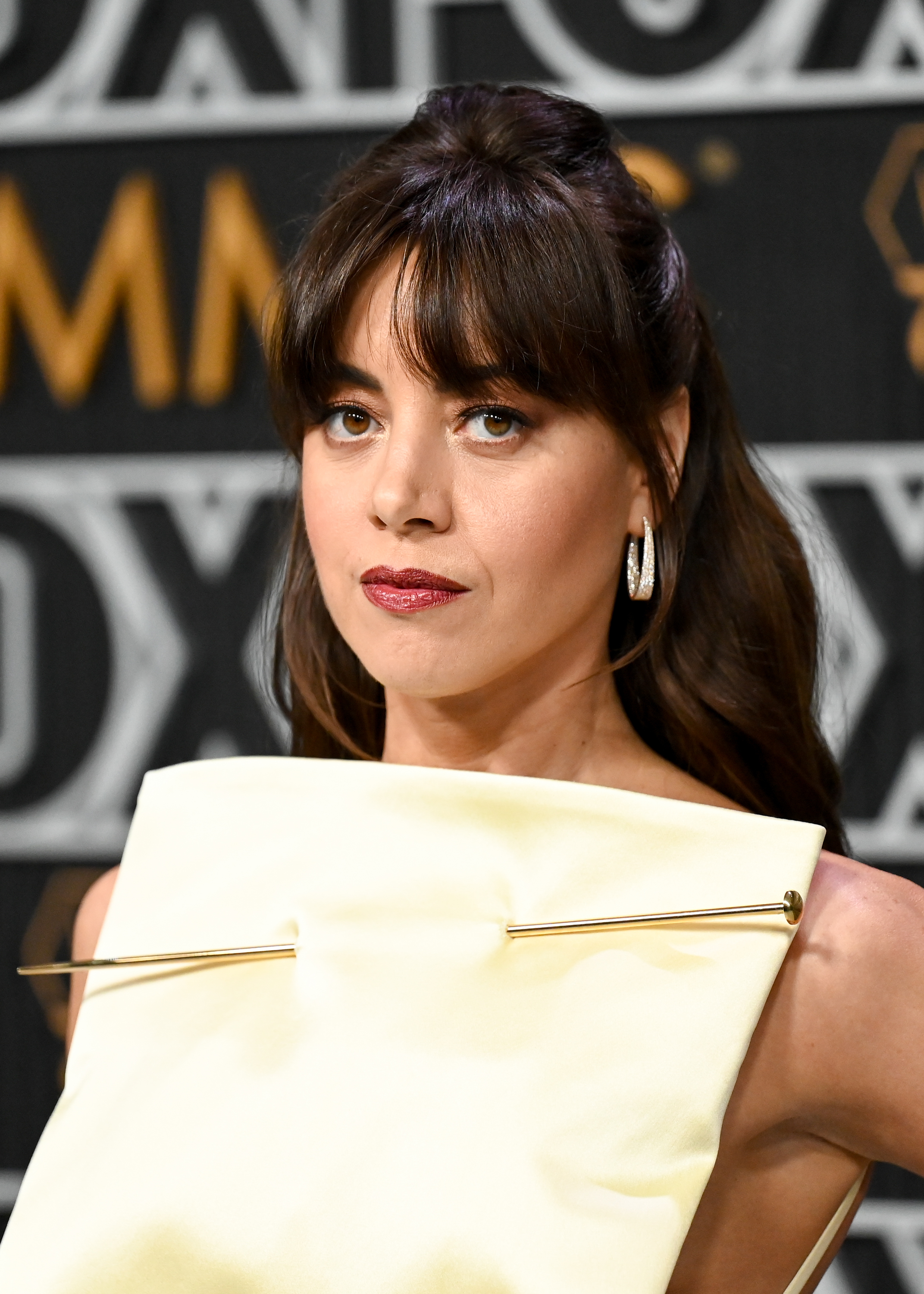 Aubrey Plaza poses on a Fox event red carpet wearing a unique, structured white outfit with large safety pins as accessories above the chest