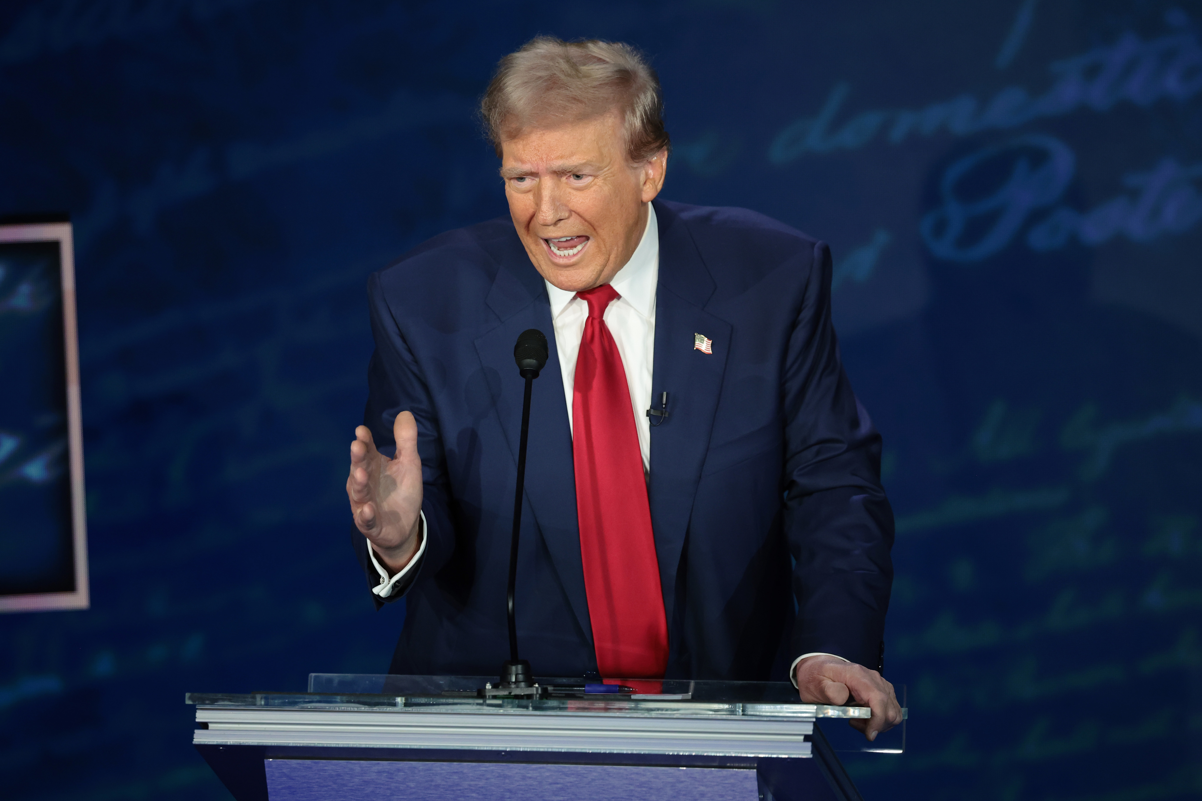 Donald Trump speaking at a podium, gesturing with his right hand and wearing a suit and red tie