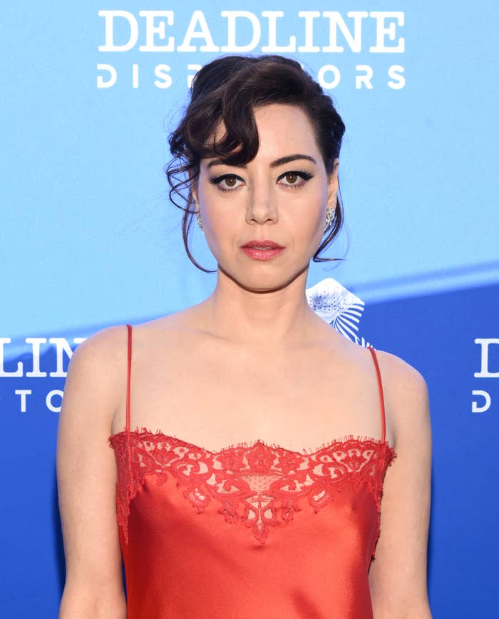 Aubrey Plaza at a Deadline event, wearing a red, lace-trimmed, spaghetti strap dress. She has an elegant updo with a side-swept bang