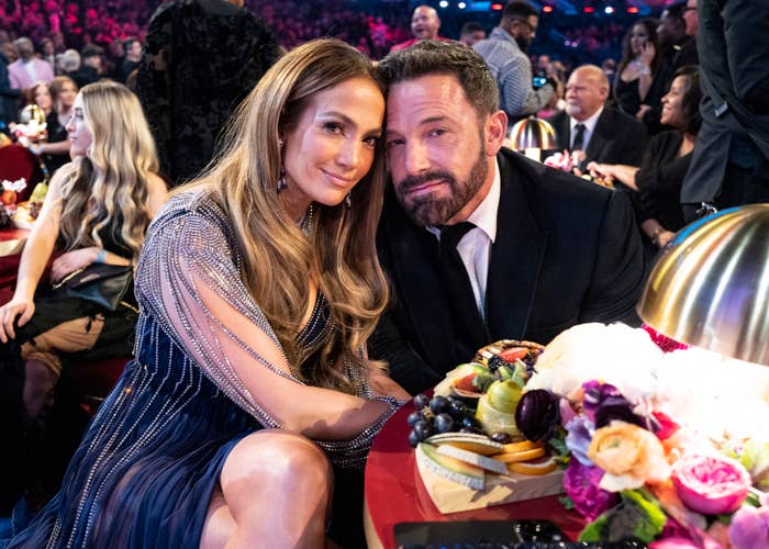 Jennifer Lopez in a beaded dress and Ben Affleck in a suit, seated close together at a formal event with others in the background