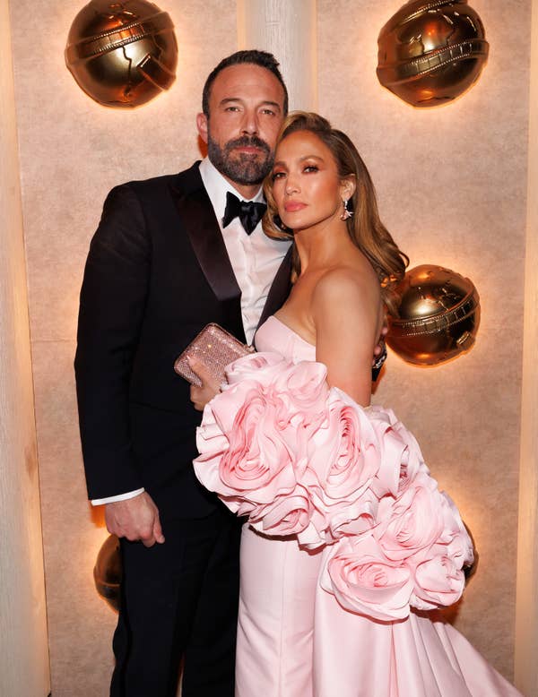 Ben Affleck in a black tuxedo and Jennifer Lopez in an elegant rose-patterned off-shoulder dress pose together at a glamorous event
