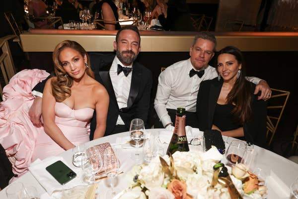 Jennifer Lopez in an elegant dress, Ben Affleck in a tuxedo, Matt Damon in a suit, and Luciana Barroso at a formal event seated at a table