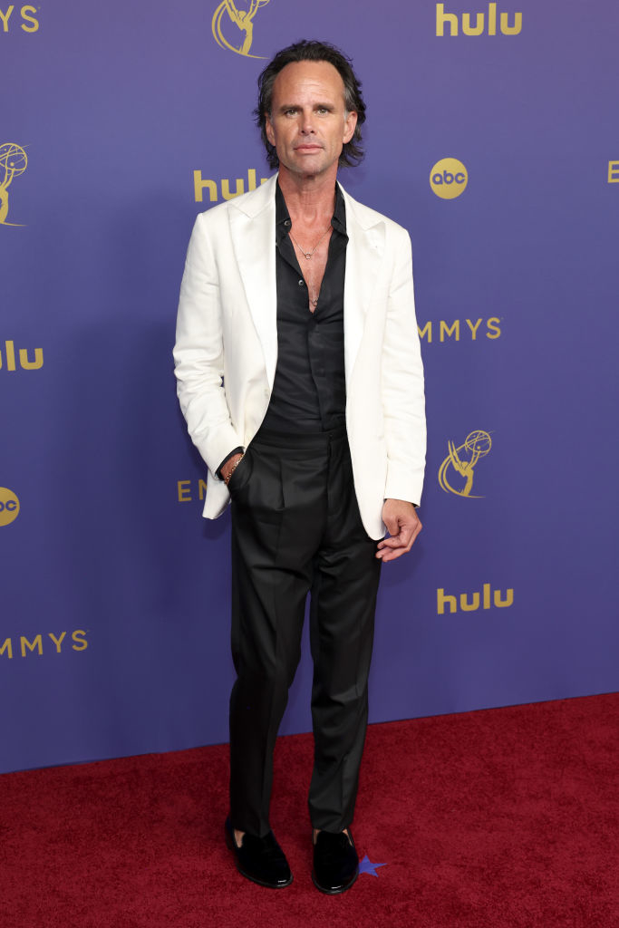 Man on red carpet wearing a white blazer, black shirt, and black pants and shoes at the Emmys