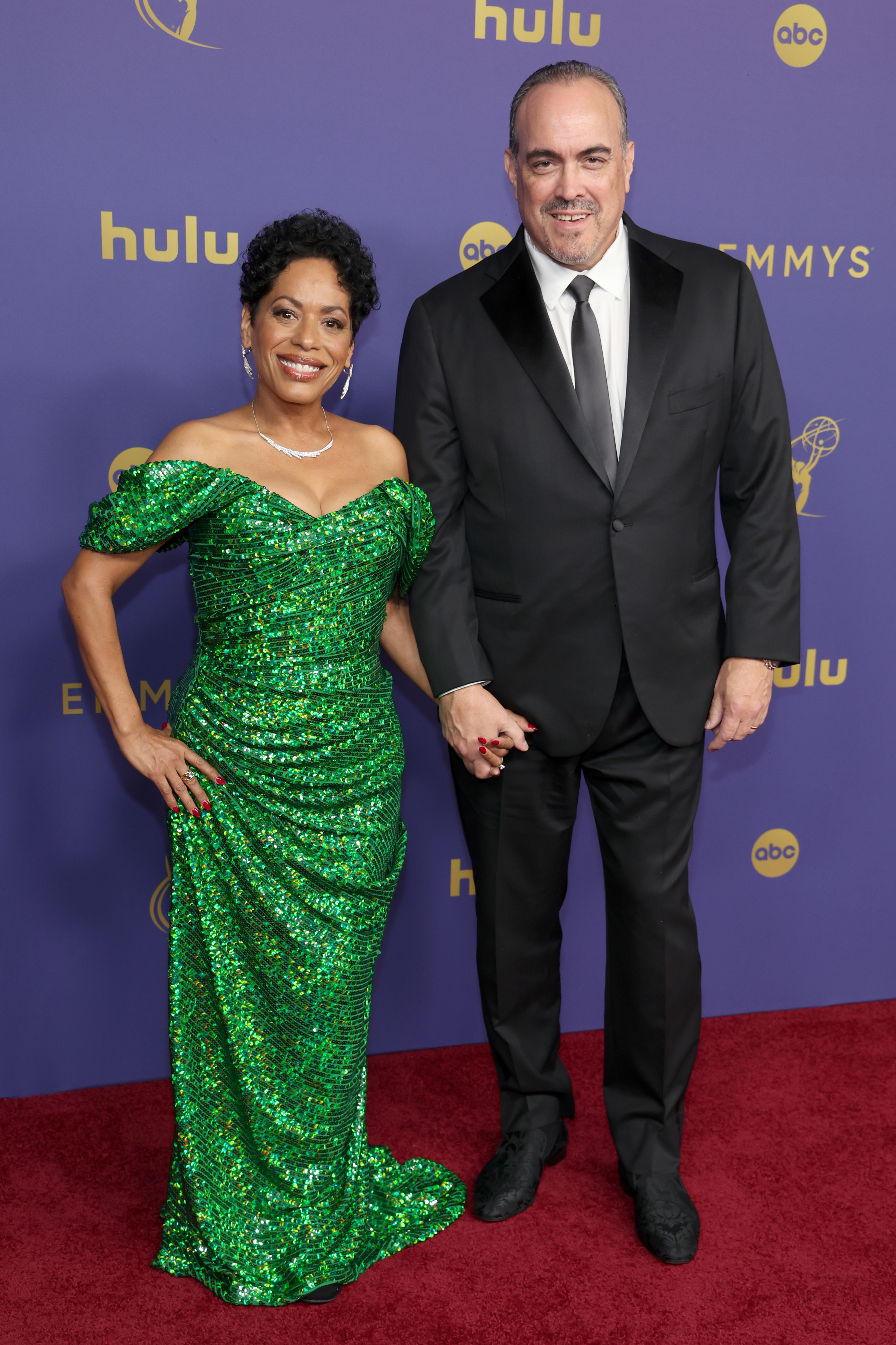 Liza Colón-Zayas in a green sequin dress and David Zayas in a black suit holding hands on the red carpet at the Emmys