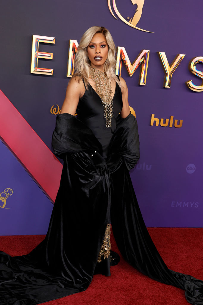 Laverne Cox poses on the red carpet at the Emmys in an elegant, flowing black gown with gold embellishments and a dramatic cape