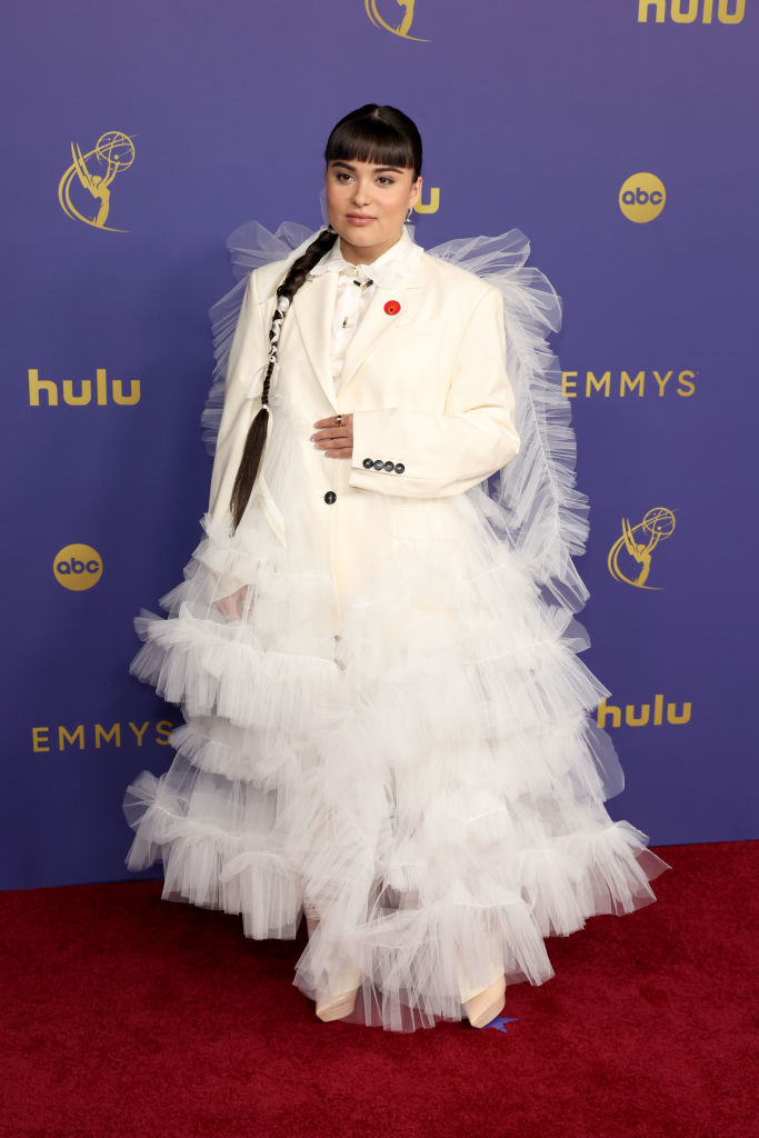 A person stands on the red carpet at the Emmys in a unique outfit consisting of a white suit jacket over a voluminous tulle skirt