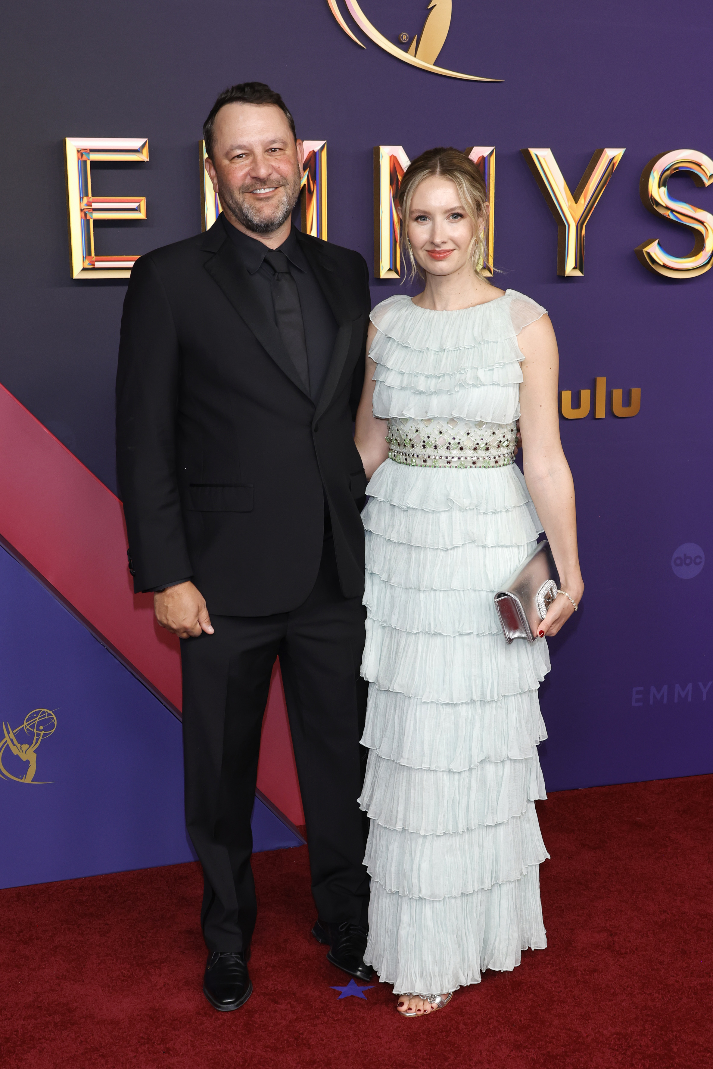 Dan Fogelman in a black suit and tie stands next to Caitlin Thompson in a tiered, sleeveless ruffled gown at the Emmy Awards