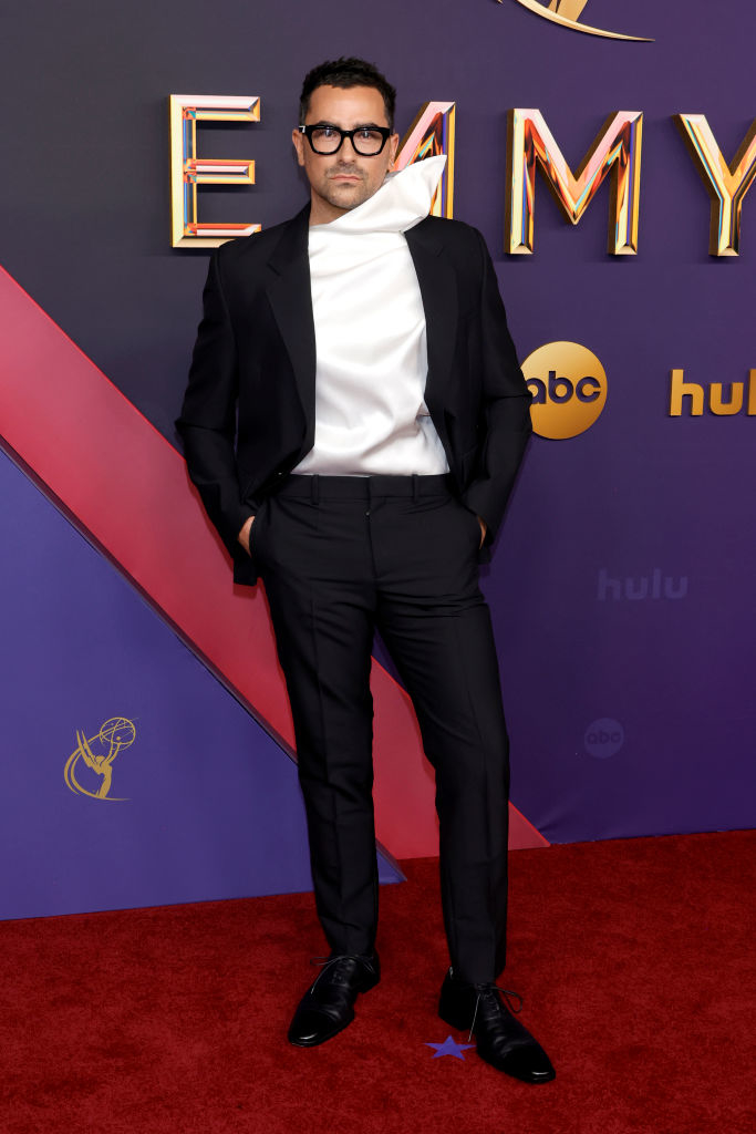 Dan Levy poses on the red carpet at the Emmys, wearing a formal suit with a unique white draped shirt, hands in pockets