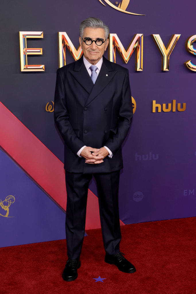 Eugene Levy at the Emmys, standing on the red carpet in a dark double-breasted suit and tie, with hands clasped