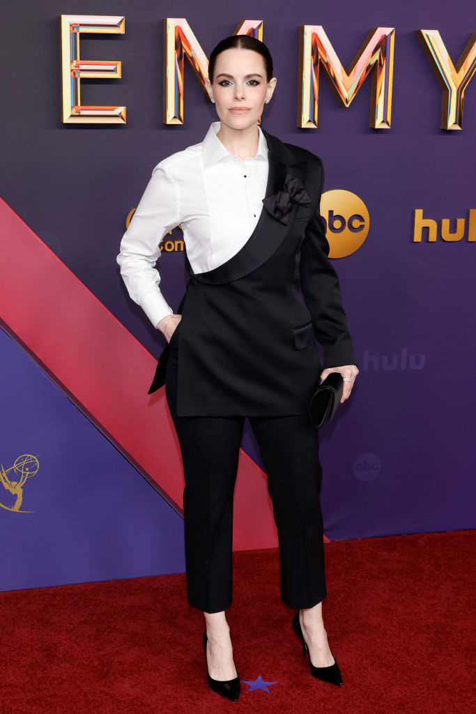 Emily Hampshire on the red carpet in a unique asymmetrical black and white suit with black pointed heels, holding a black clutch at the Emmy Awards
