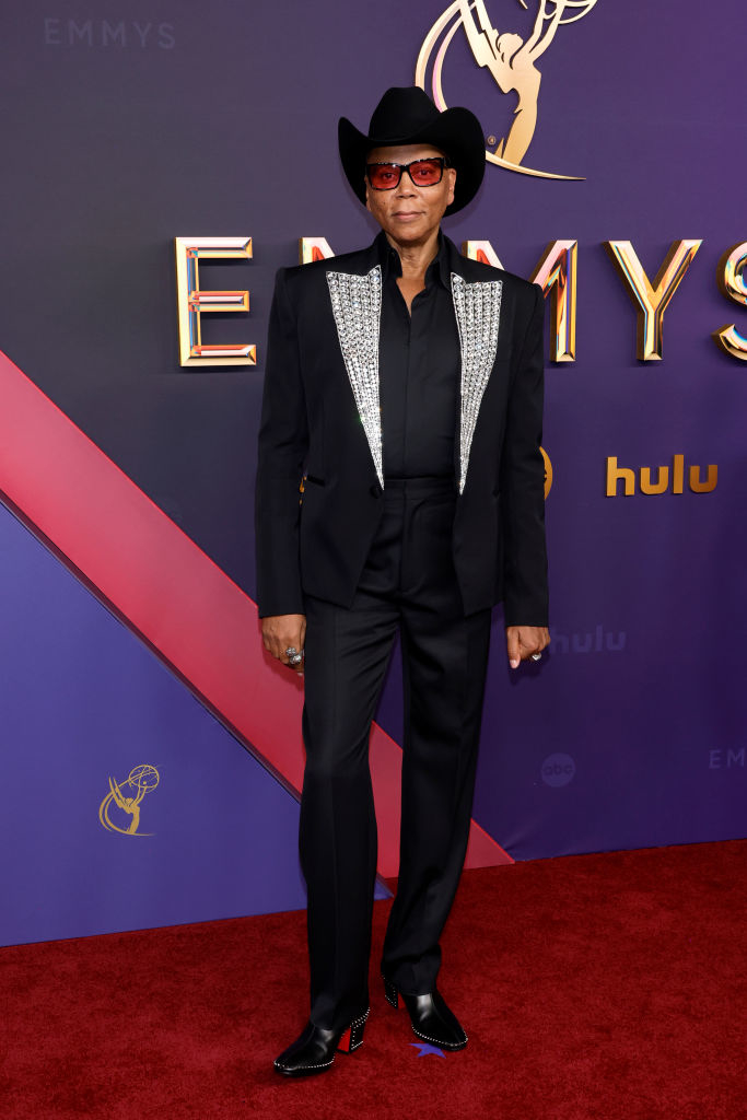 RuPaul on the red carpet wearing a black suit with jeweled lapels, black shirt, black hat, and red sunglasses, at an Emmy Awards event