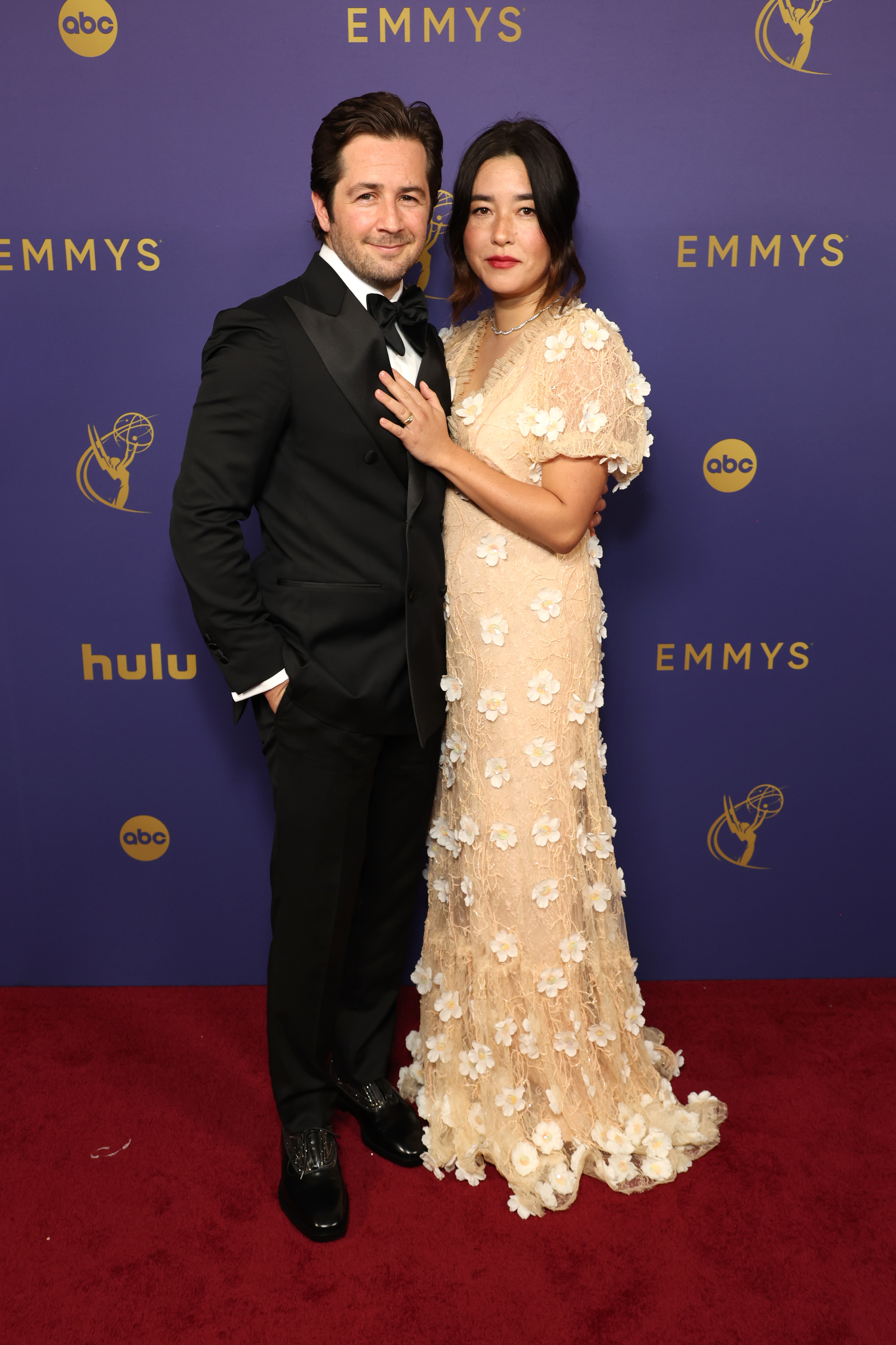 Michael Angarano and Maya Erskine posing on the Emmys red carpet. Michael is in a black tuxedo, and Maya is in a floral, floor-length gown
