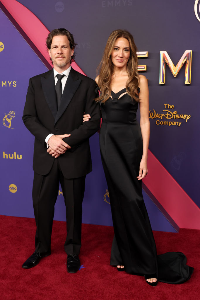 Man and woman at the Emmys. The man wears a black tuxedo, while the woman is in a sleek black gown. They pose together on the red carpet