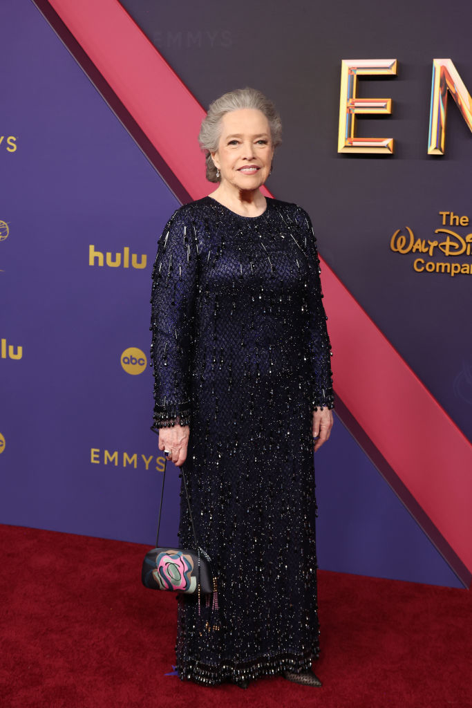 Kathy Bates on the red carpet at the Emmy Awards, wearing a long-sleeved, black dress adorned with sequin details, holding a small handbag with a colorful design