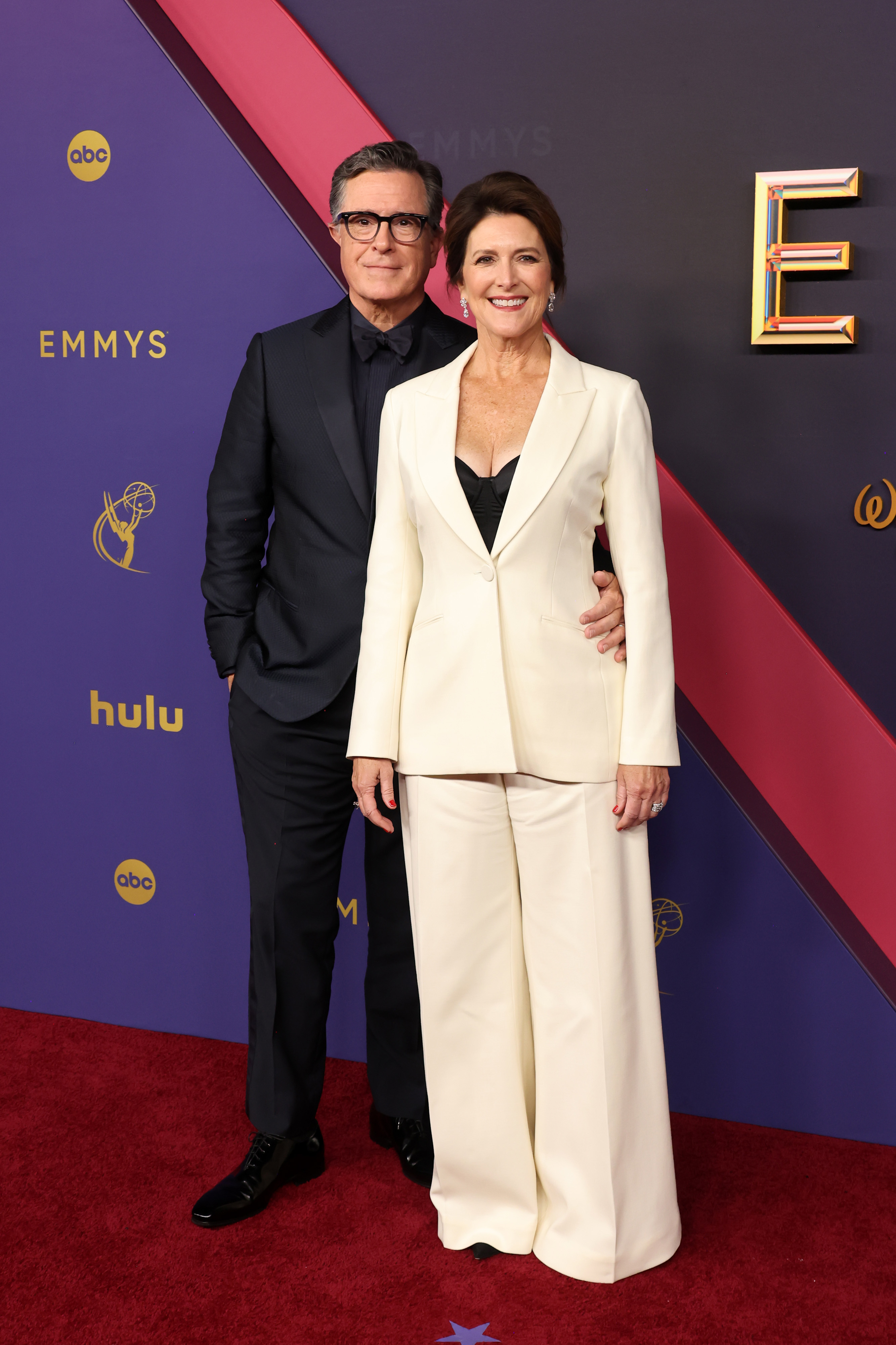 Stephen Colbert and Evelyn McGee-Colbert on the red carpet, with Stephen in a dark suit and Evelyn in a white pantsuit with a black top at the Emmy Awards