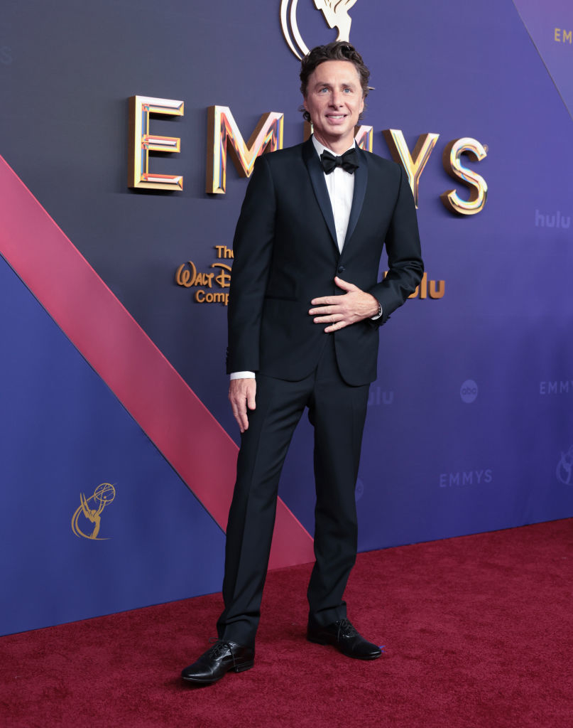 Zach Braff stands on the red carpet at the Emmys, wearing a classic black tuxedo with a black bow tie. He smiles and poses with one hand on his stomach