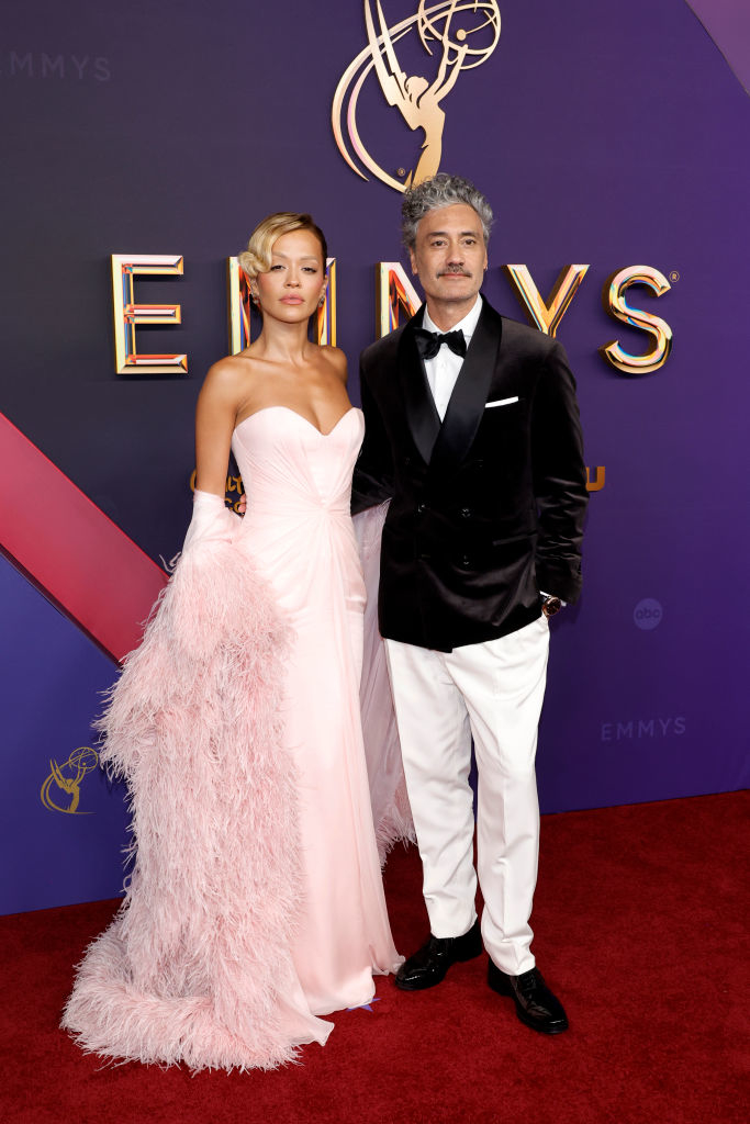 Rita Ora in a strapless gown with feathered details, and Taika Waititi in a tuxedo at a red carpet event