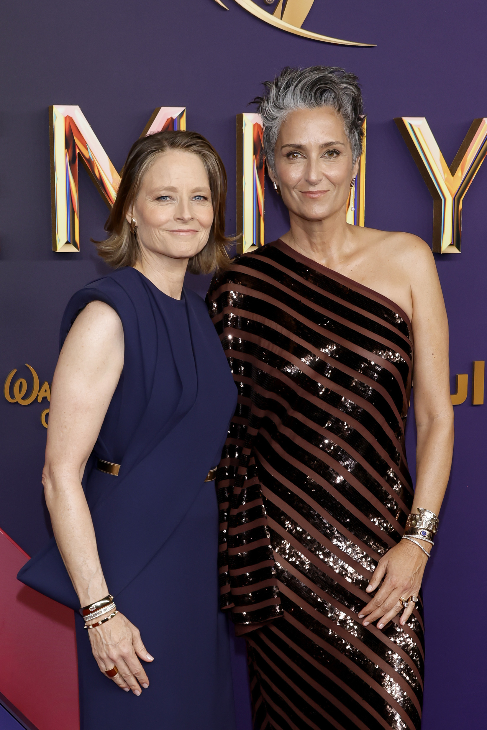 Jodie Foster and Alexandra Hedison on the red carpet. Foster wears a stylish navy dress, and Hedison is in a striking one-shoulder sequined dress with striped pattern