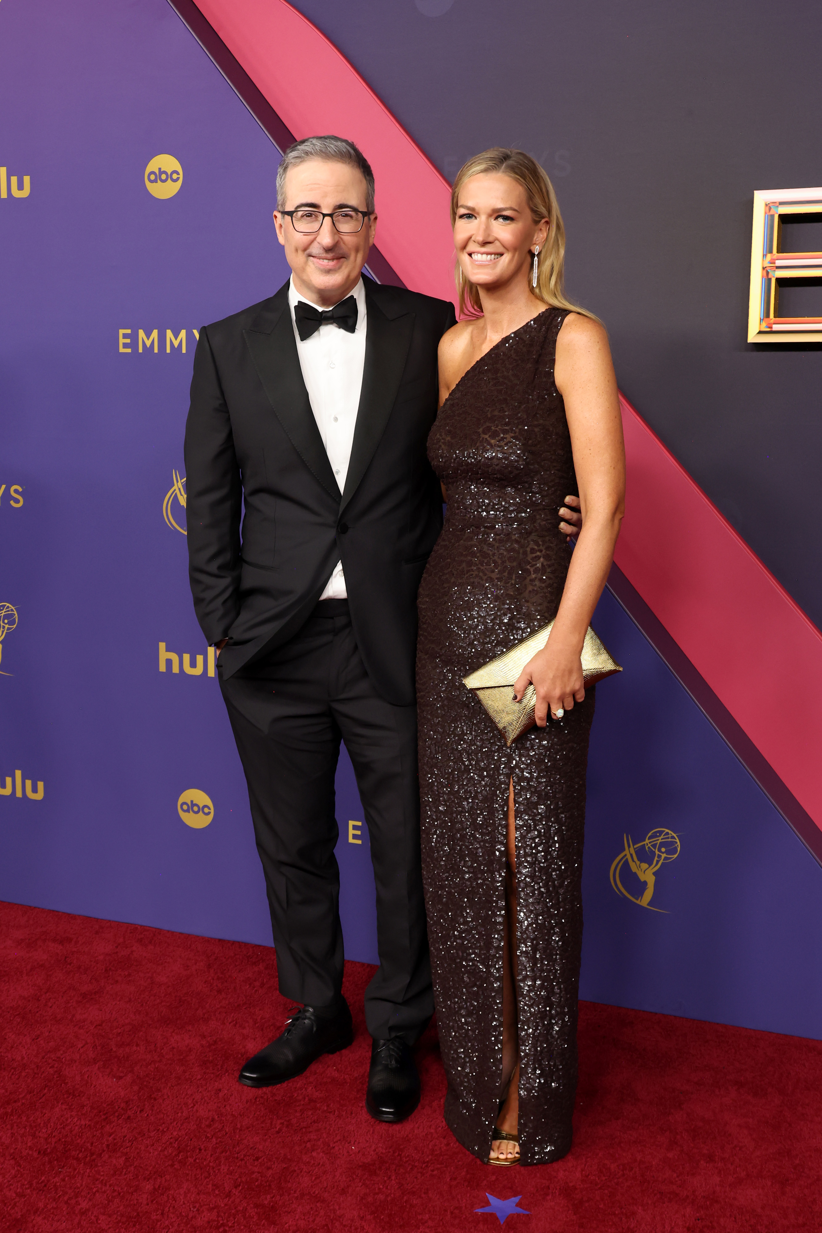 John Oliver in a tuxedo and Kate Norley in a sequined one-shoulder gown, pose together on the red carpet at the Emmys