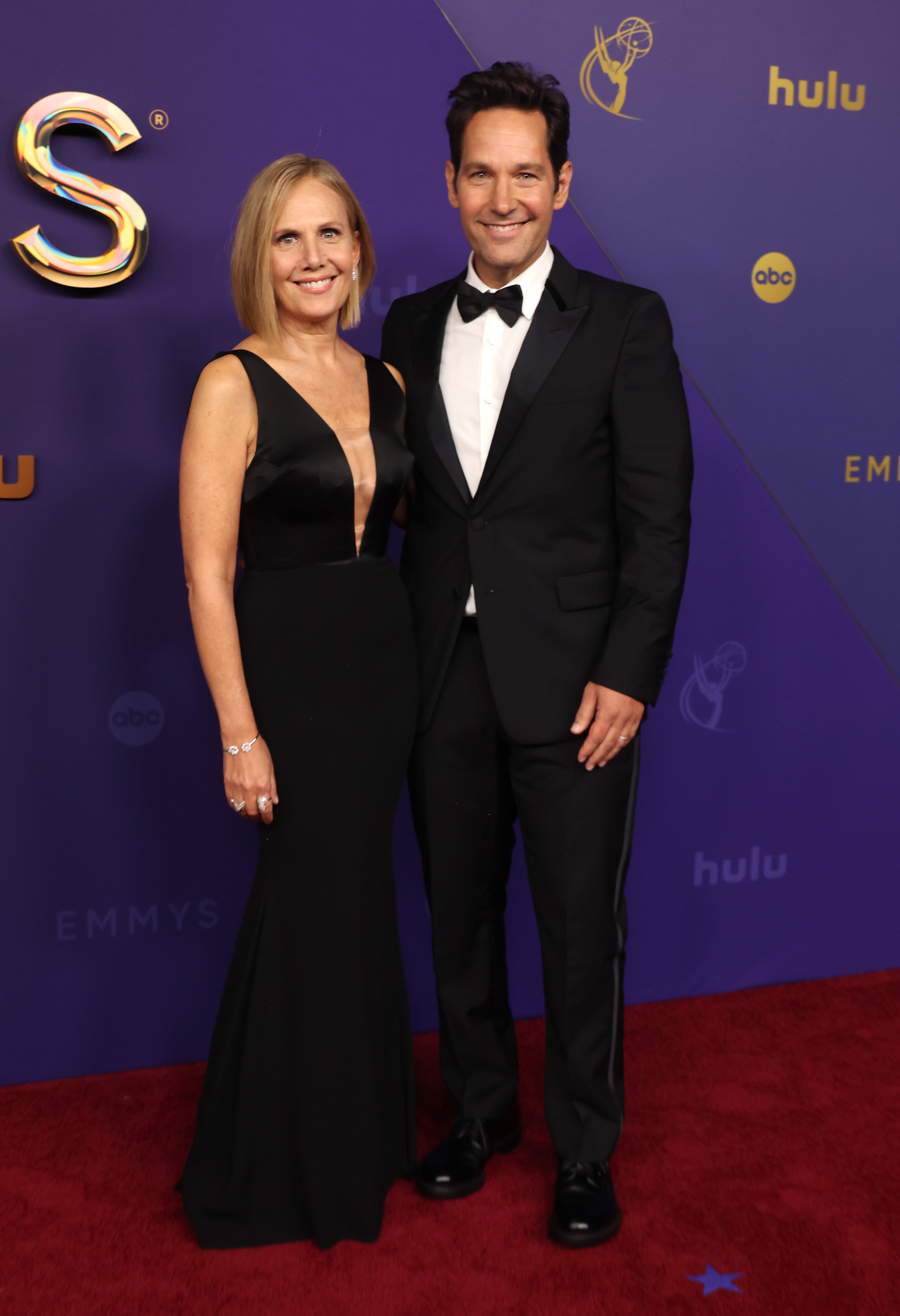 Julie Yaeger and Paul Rudd stand on the red carpet. Julie wears a sleeveless black gown, and Paul wears a black tuxedo with a black bow tie