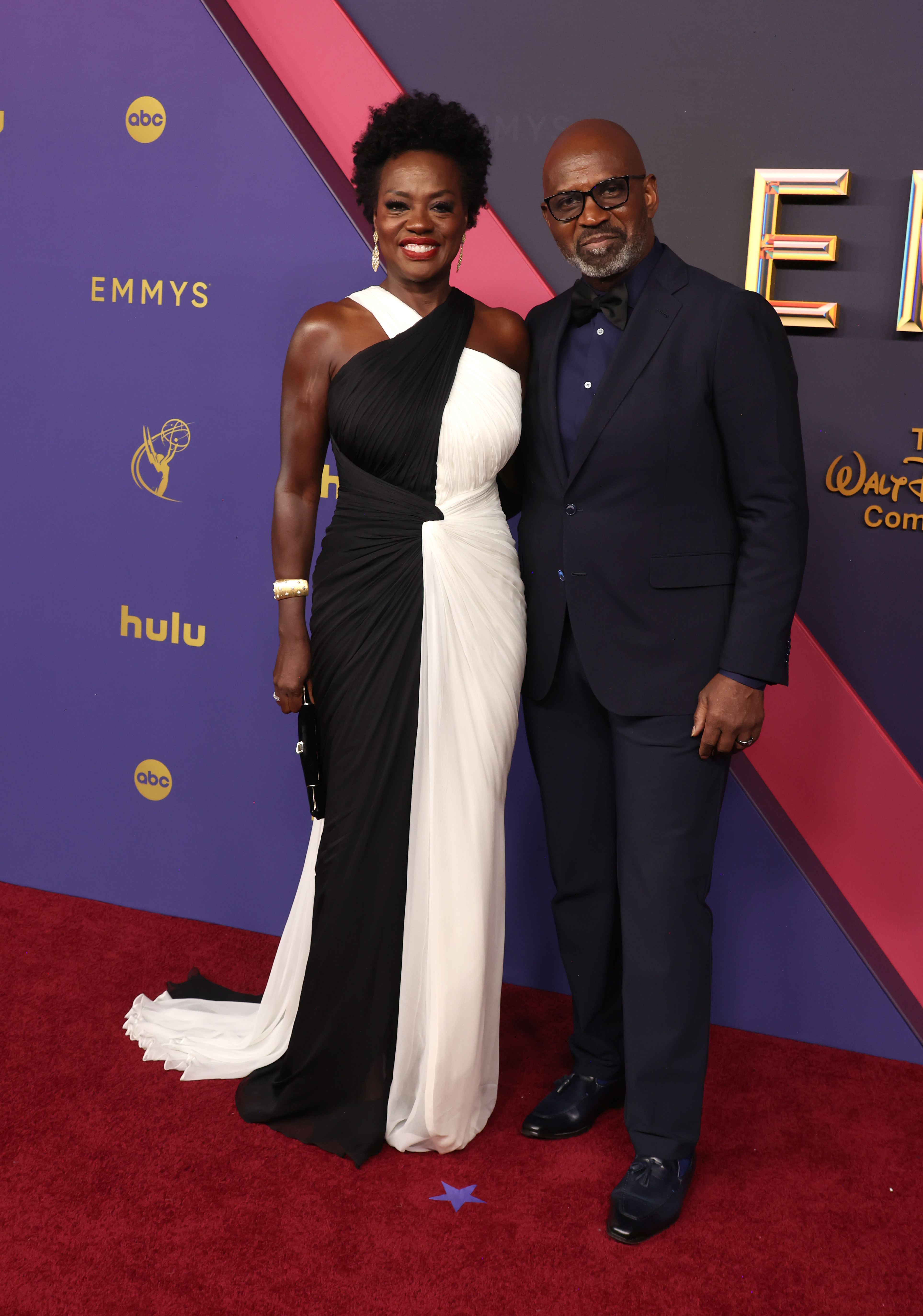 Viola Davis and Julius Tennon stand together on the red carpet at the Emmys. Viola wears an elegant monochrome gown, while Julius dons a sleek dark suit and glasses
