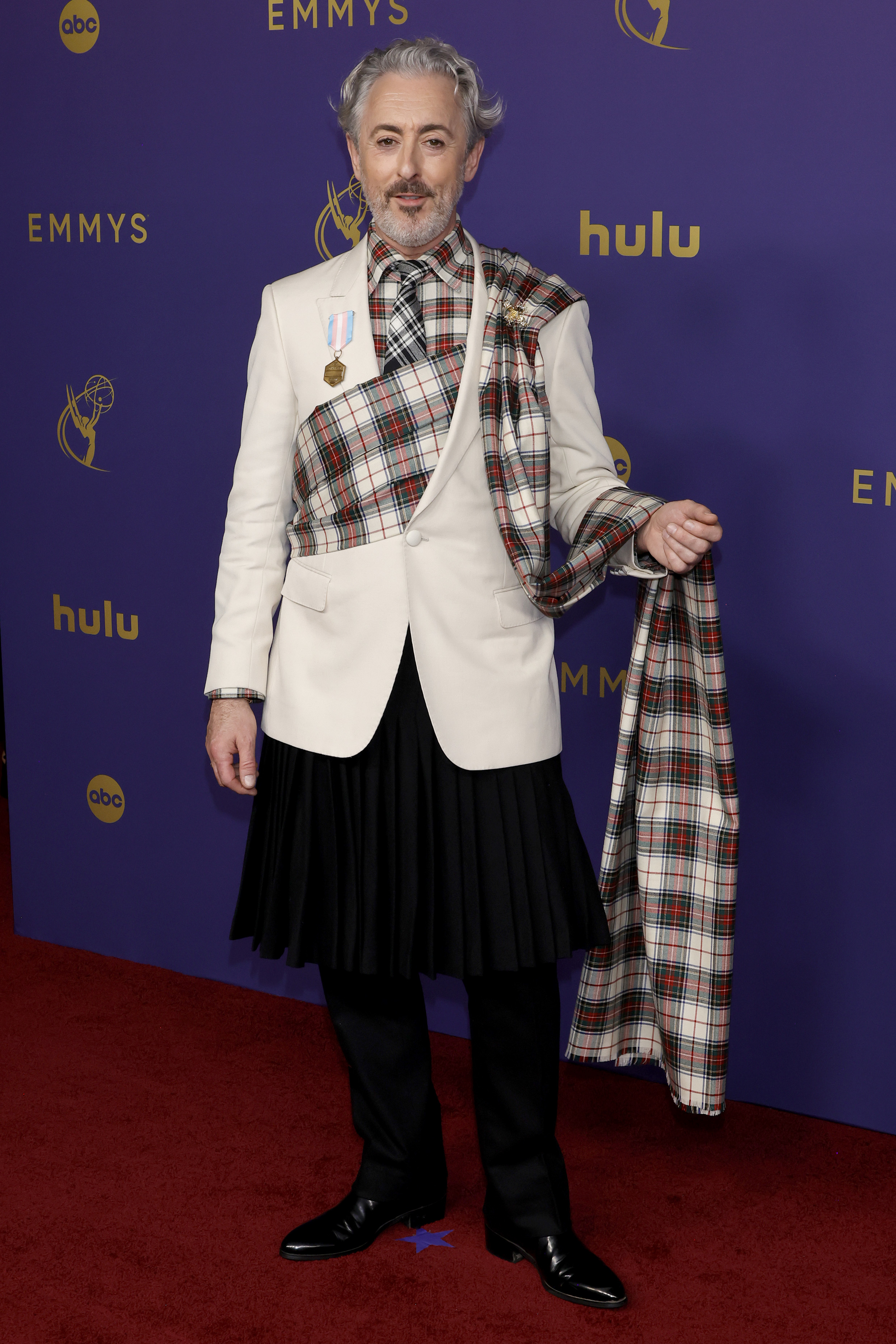 Alan Cumming is on the red carpet at the Emmys, wearing a jacket, kilt, and a plaid sash