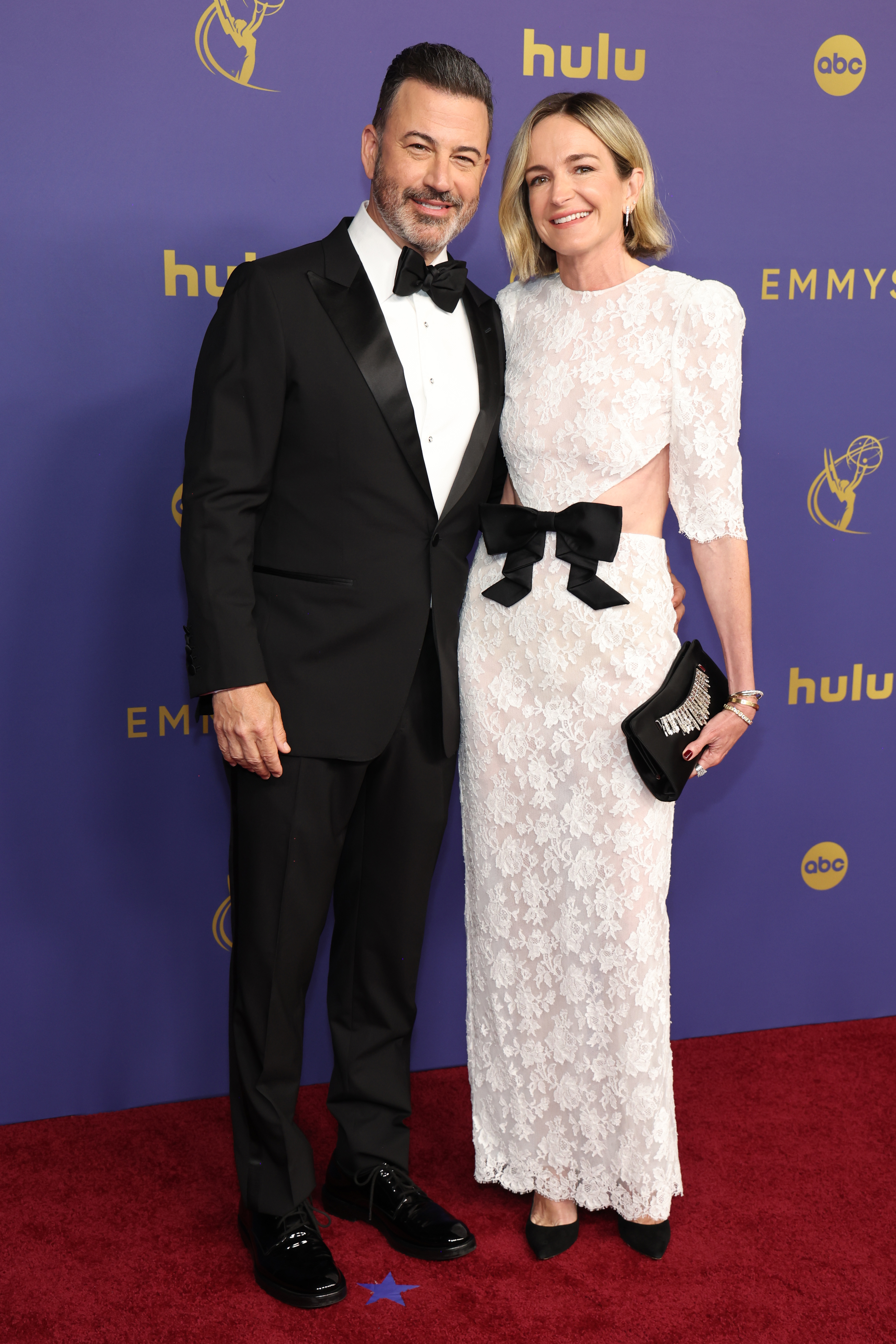 Jimmy Kimmel in a black tuxedo and Molly McNearney in a white lace dress with cutouts and a black bow, at the Emmy Awards red carpet