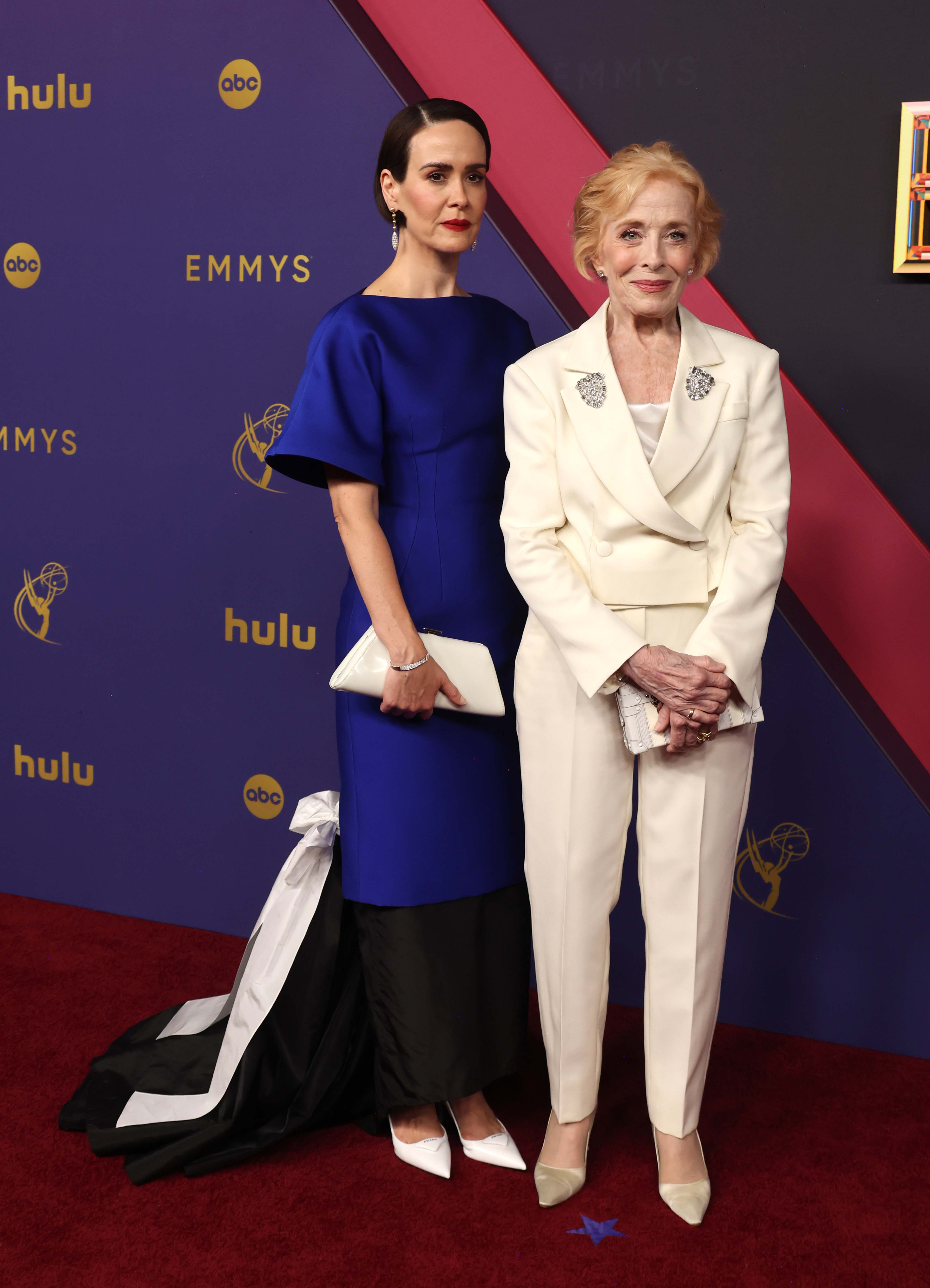 Sarah Paulson in a blue dress with a black and white train, standing beside Holland Taylor in a white suit on the red carpet at the Emmys